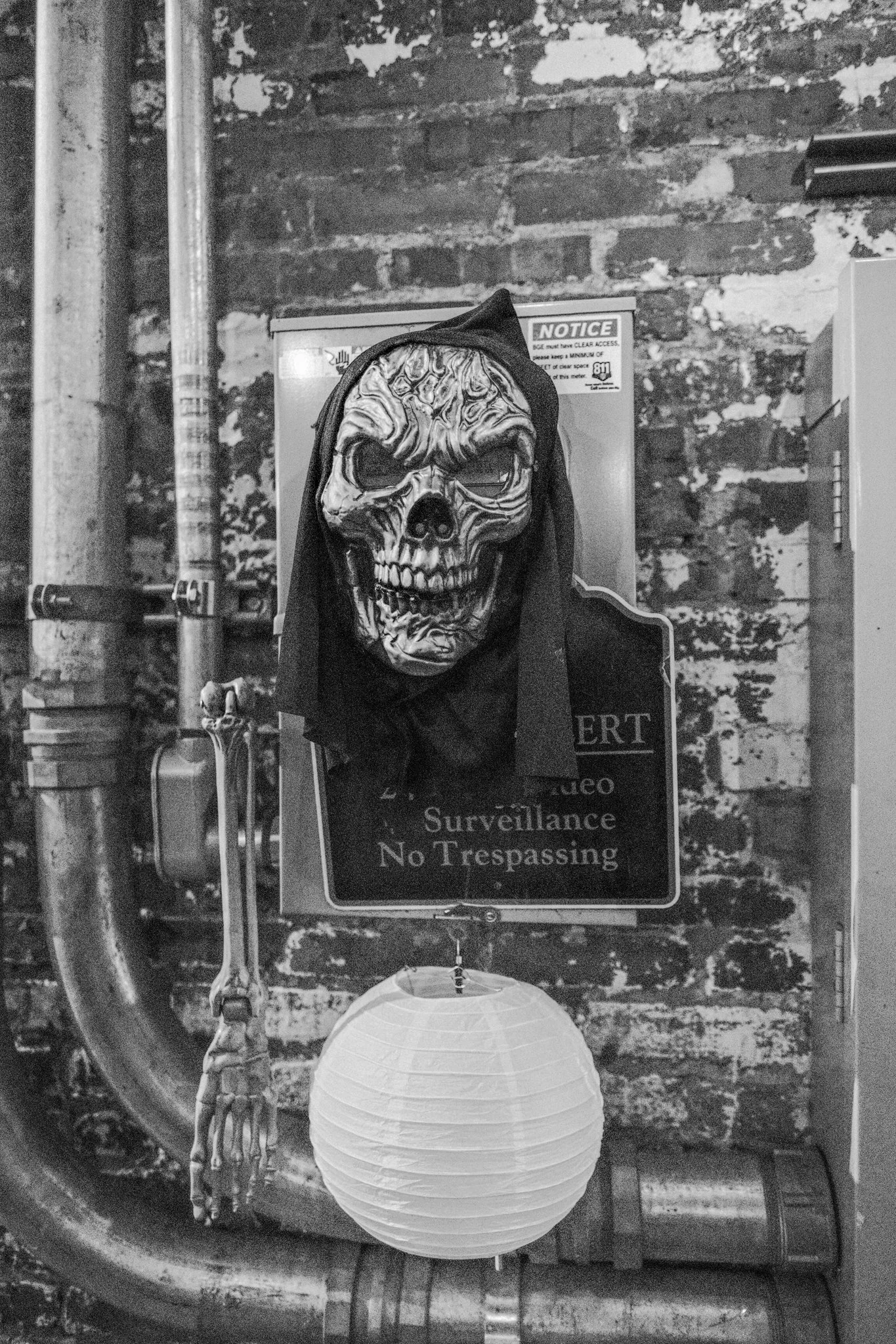 A skeleton wearing a skull mask and black hood hangs on a wall, with a paper lantern hanging below and pipes nearby.