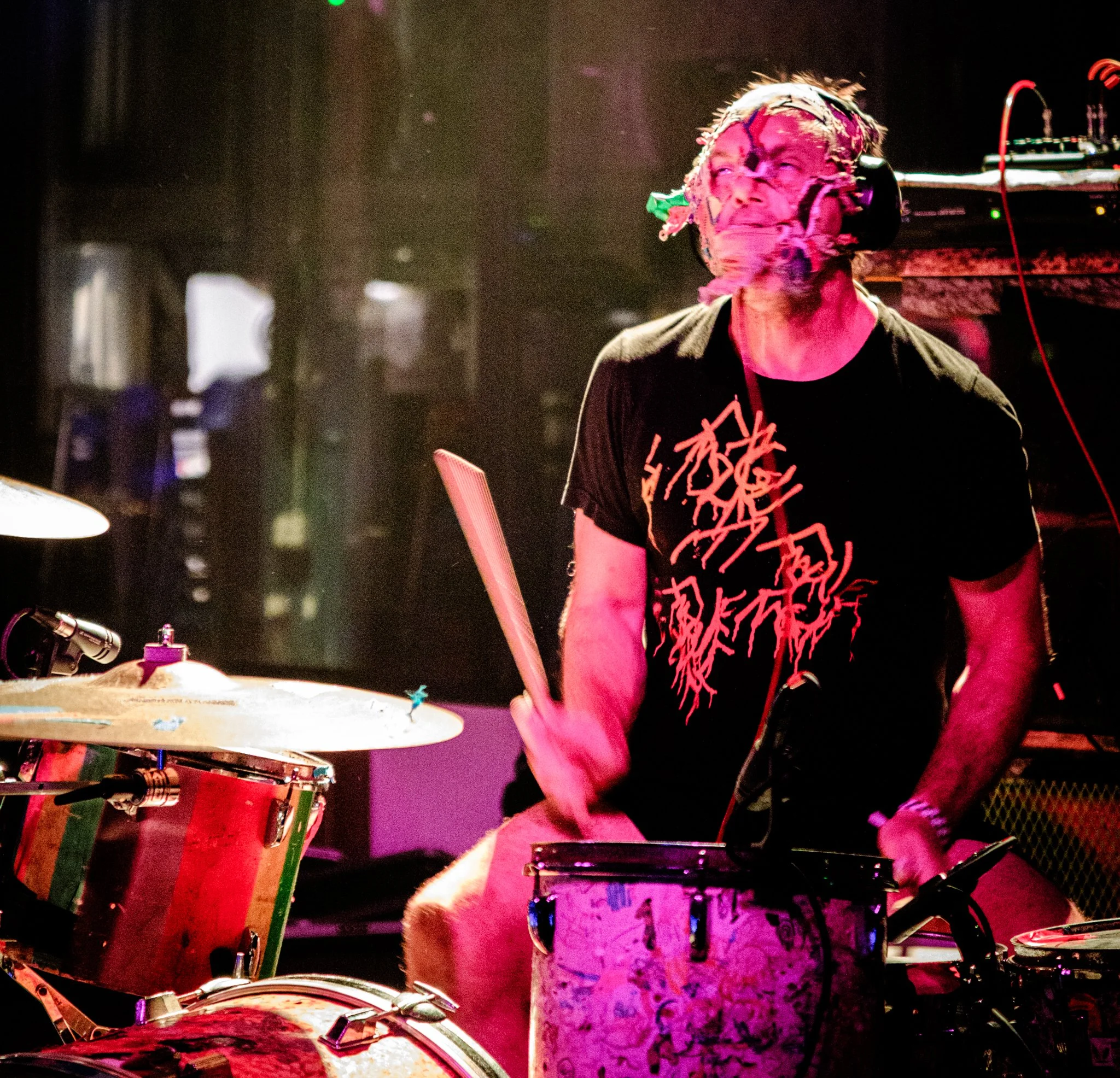 A drummer wearing a black T-shirt with red writing, playing drums on stage under colorful lighting.