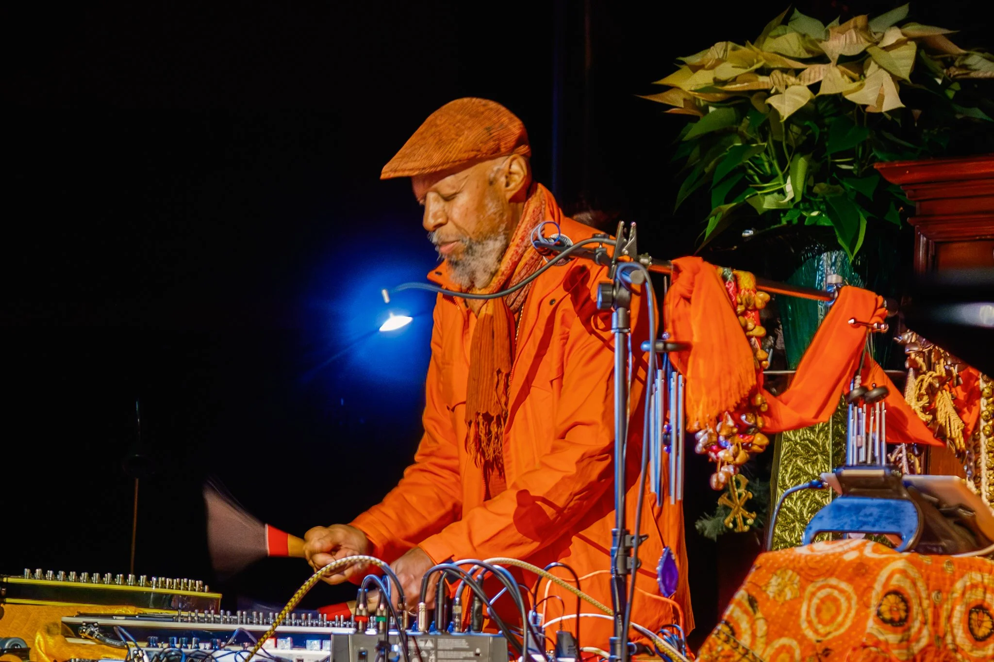 An elderly man with a gray beard and closed eyes, dressed in bright orange clothing, is DJing behind a table with electronic music equipment, set against a dark background with a plant and decorative cloth.