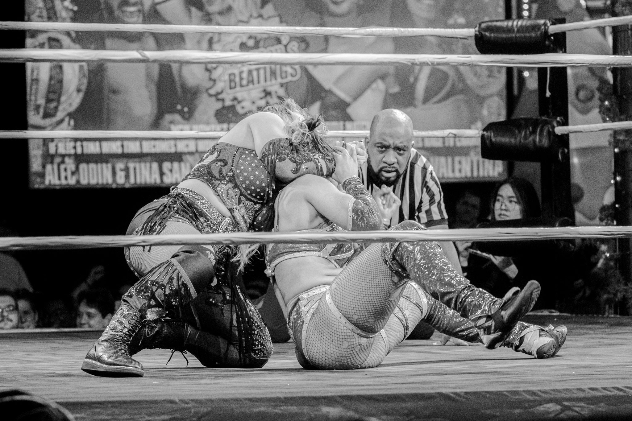 Two female wrestlers on the mat in a wrestling ring, one pinning the other, with a referee observing closely, and a woman and another person in the background watching.