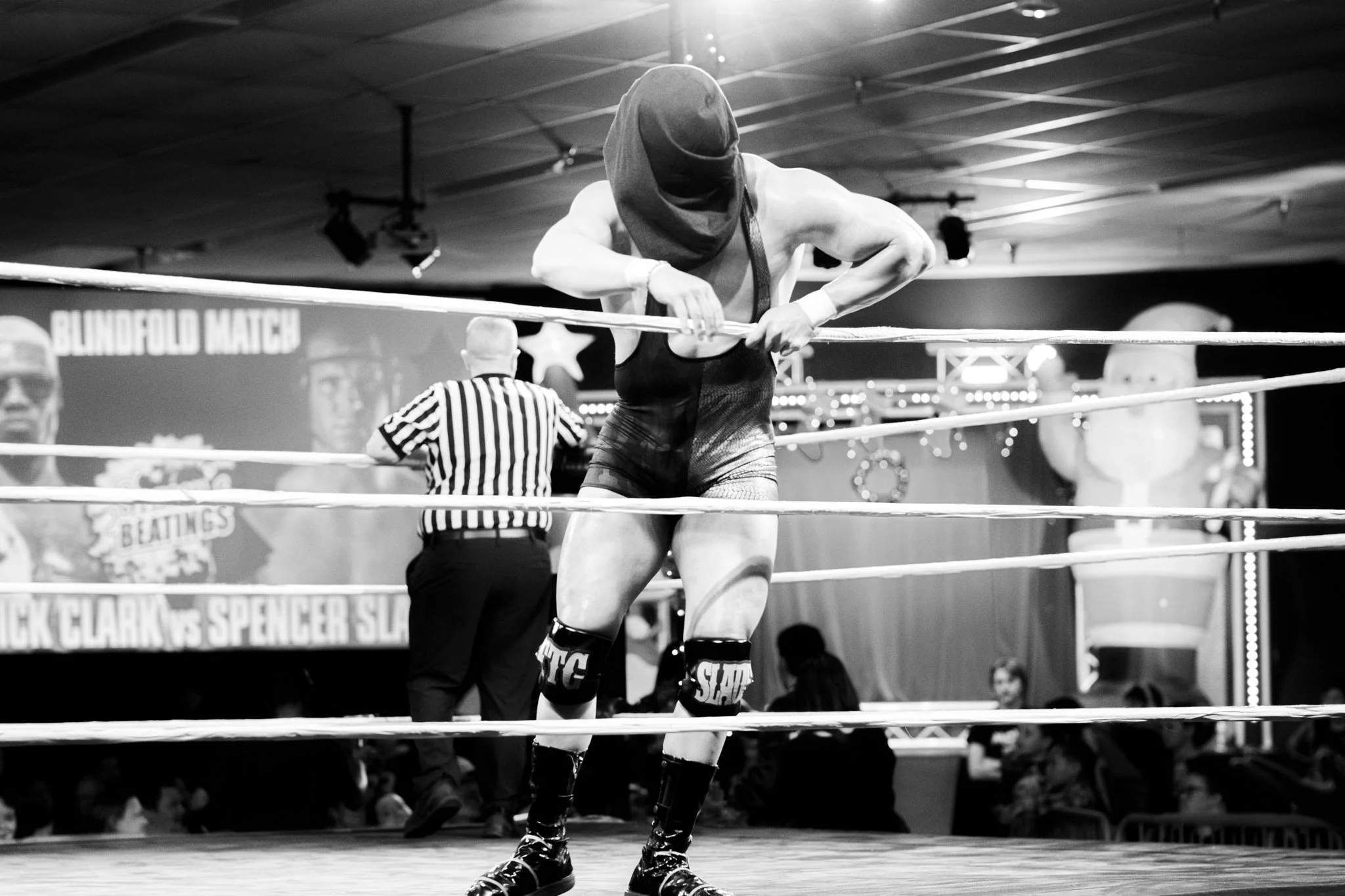 A wrestler wearing a face mask and sports attire is adjusting the wrestling ring ropes during a match. A referee and audience can be seen in the background.