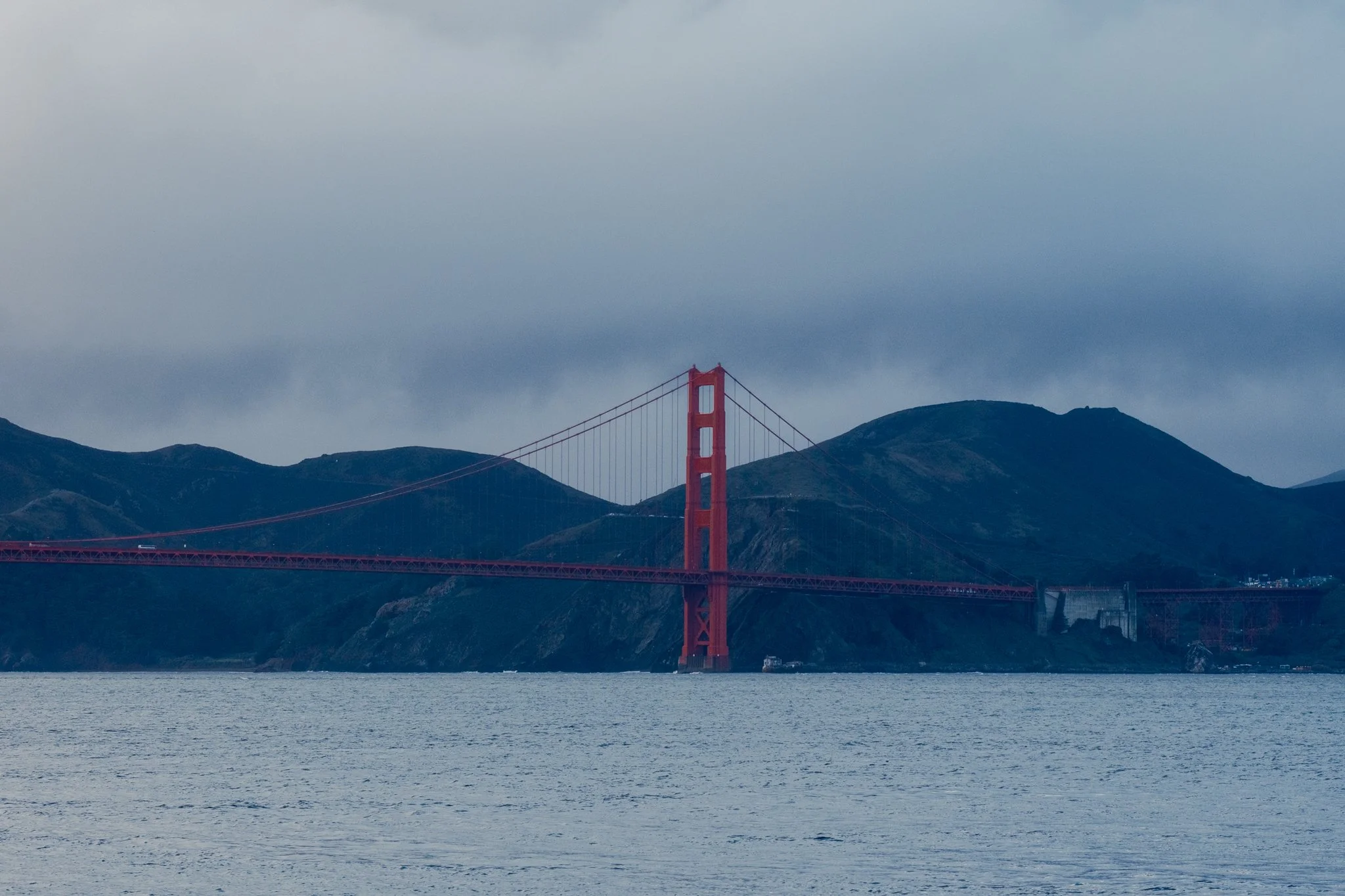 The Golden Gate Bridge in San Francisco, California, with mountains in the background and water in the foreground under a cloudy sky.