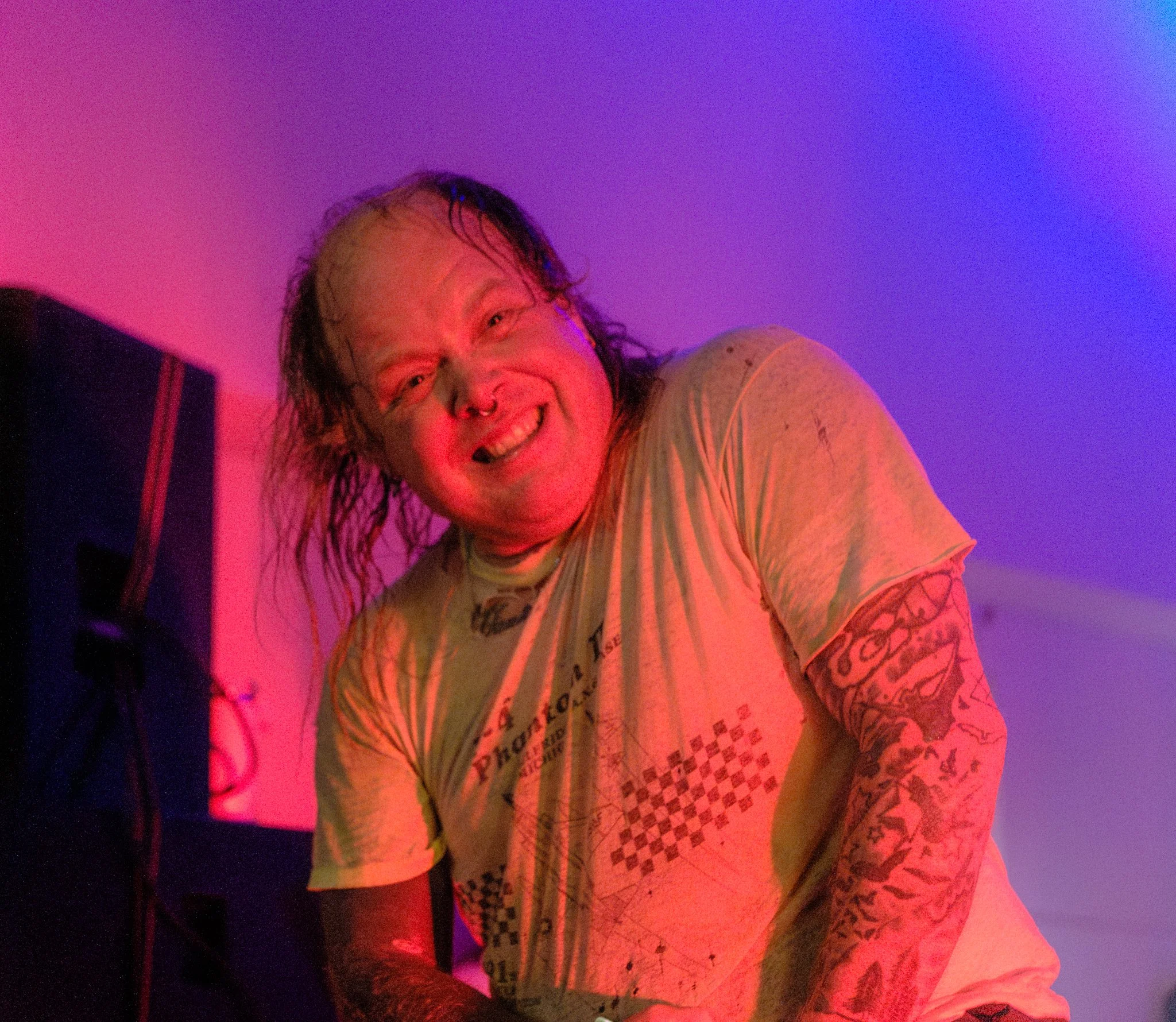 A person with long hair and tattoos smiling in colorful lighting.