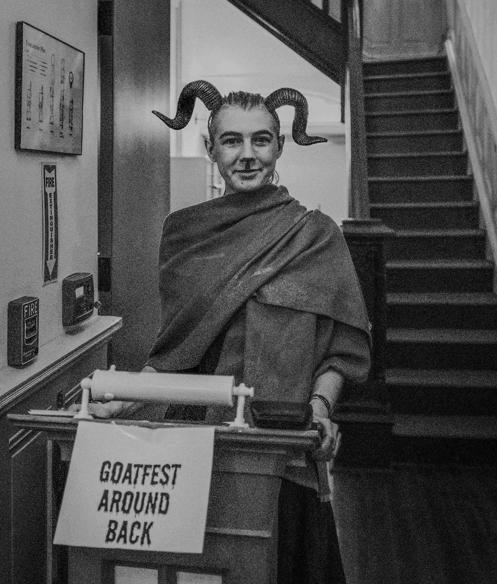 Person with ram horns headband and cape at event table with sign that reads 'Goatfest Around Back', standing indoors near stairs.