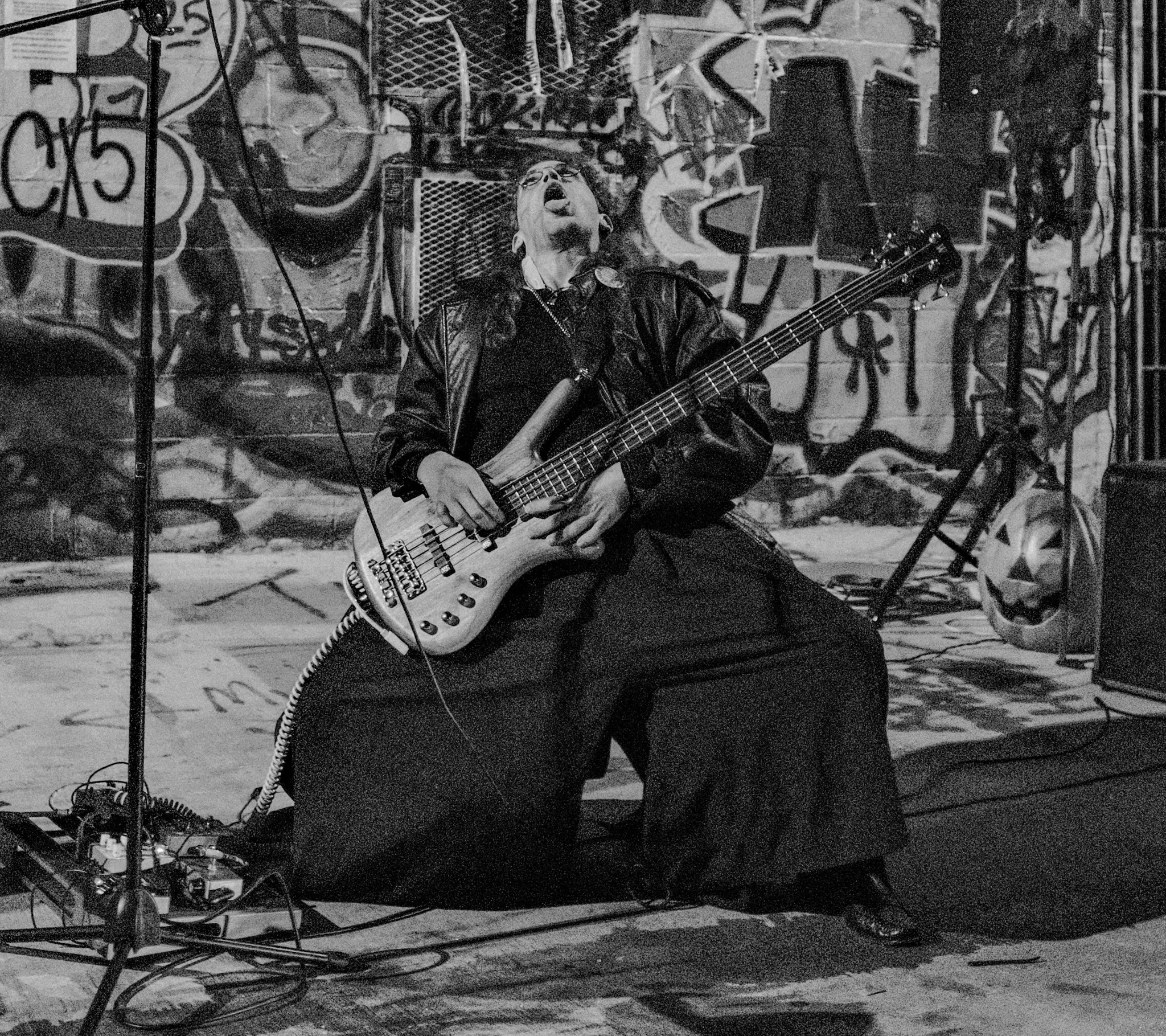 A musician is playing a bass guitar on stage, sitting on a stool, with a graffiti-covered wall behind him. The musician has long hair, wears glasses, and is dressed in dark clothing, including a leather jacket. The scene is in black and white.
