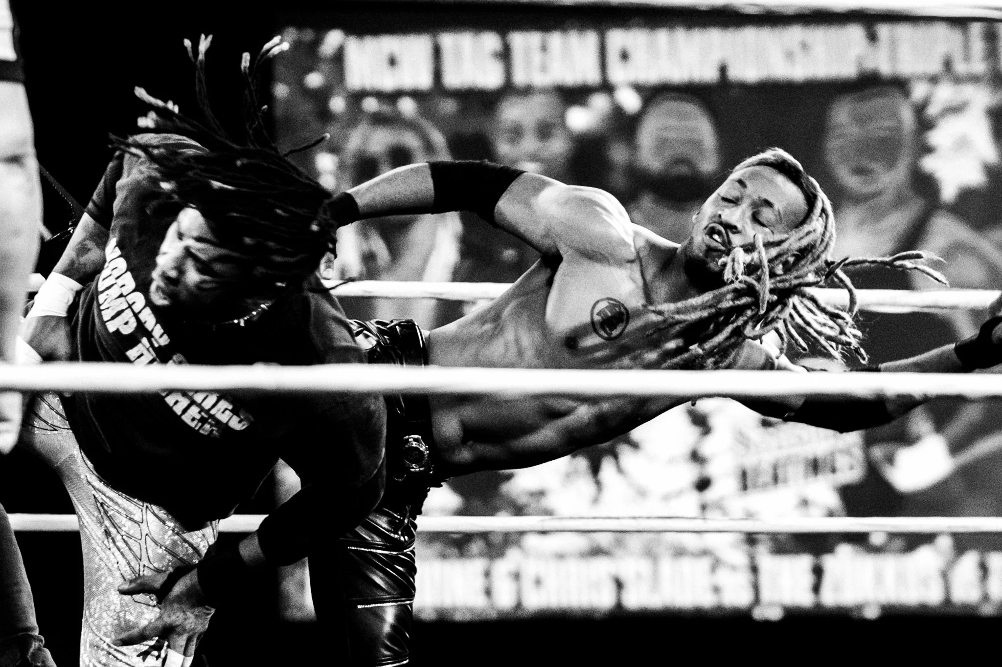 A black and white photo of two wrestlers in a wrestling ring, one with long dreadlocks and the other with shorter hair, engaging in a match with ropes visible in the foreground.
