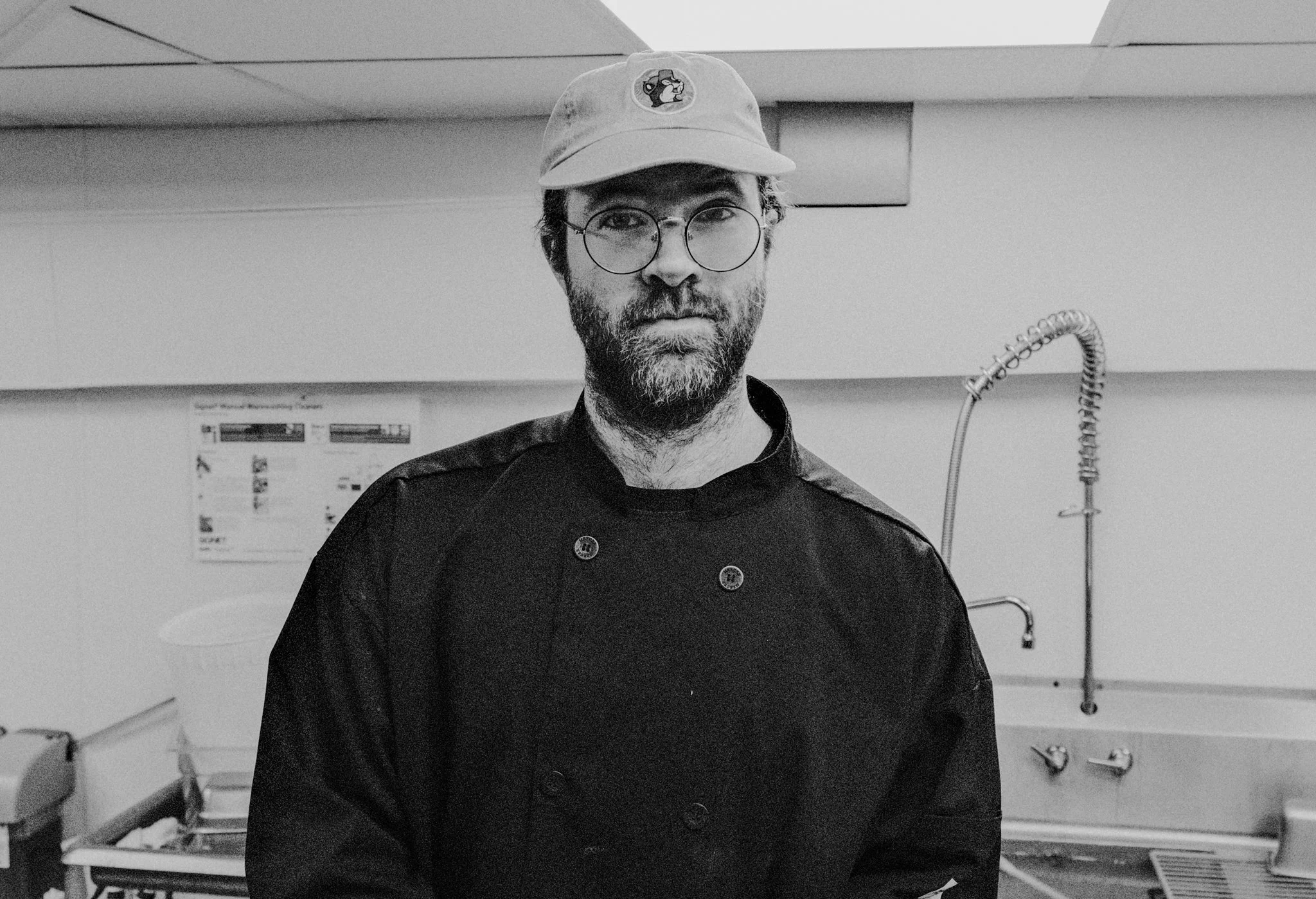 A man with glasses and a beard, wearing a chef's coat and a baseball cap with a mascot logo, standing in a kitchen.