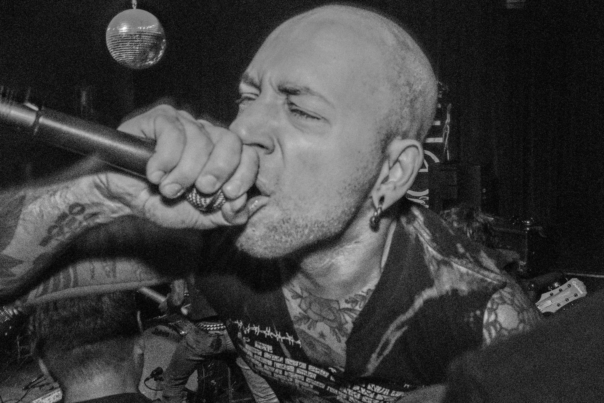 A man with a shaved head and tattoos sings into a microphone in a dark room with a disco ball overhead.