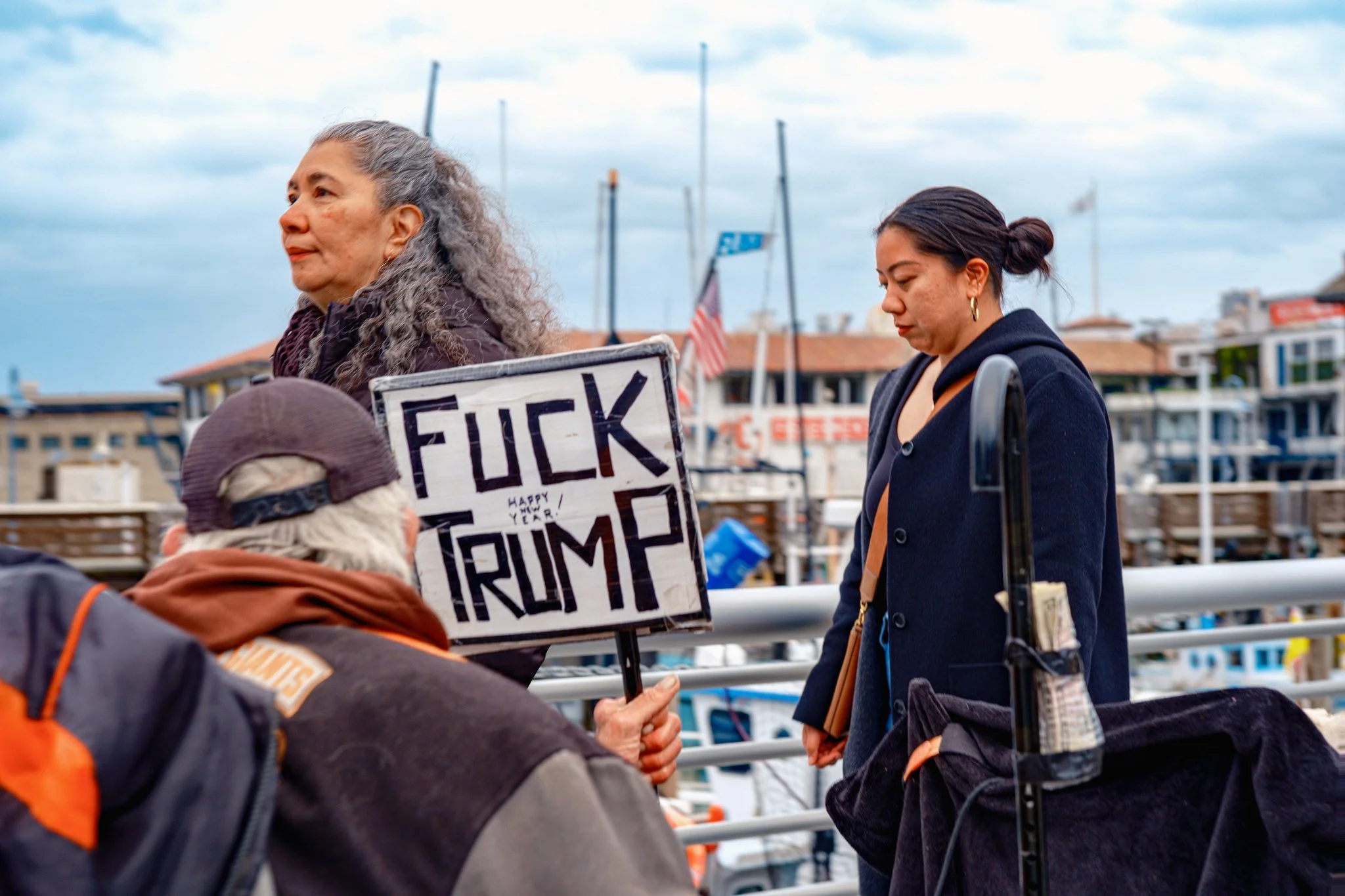 A person holding a protest sign that reads 'FUCK TRUMP' with a small 'Happy New Year!' note, standing near two women by a marina with boats and buildings in the background.