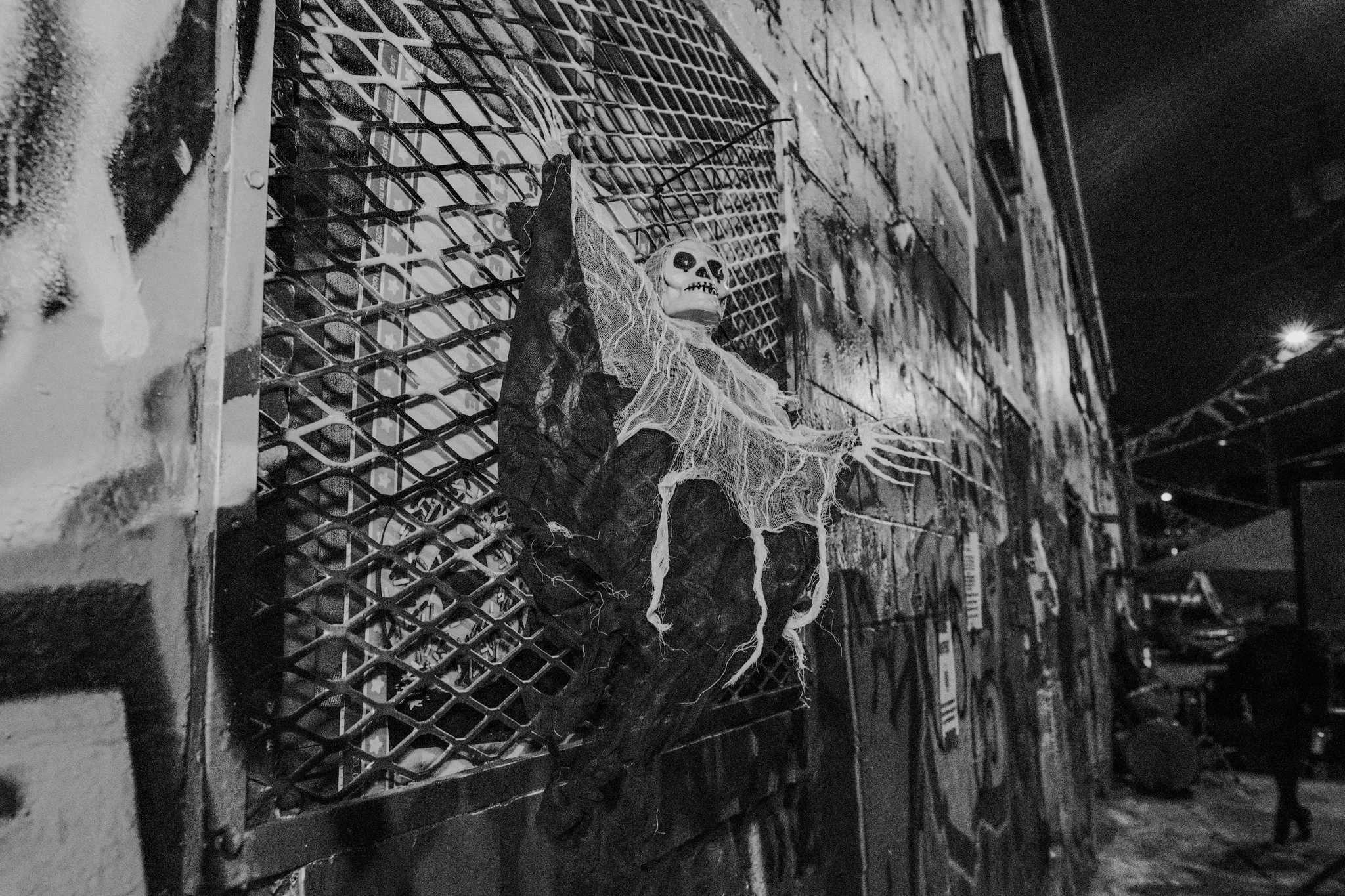 Halloween decoration of a skeleton with long hair and tattered clothing hanging on a metal grate on a brick wall, in a dimly lit outdoor area.