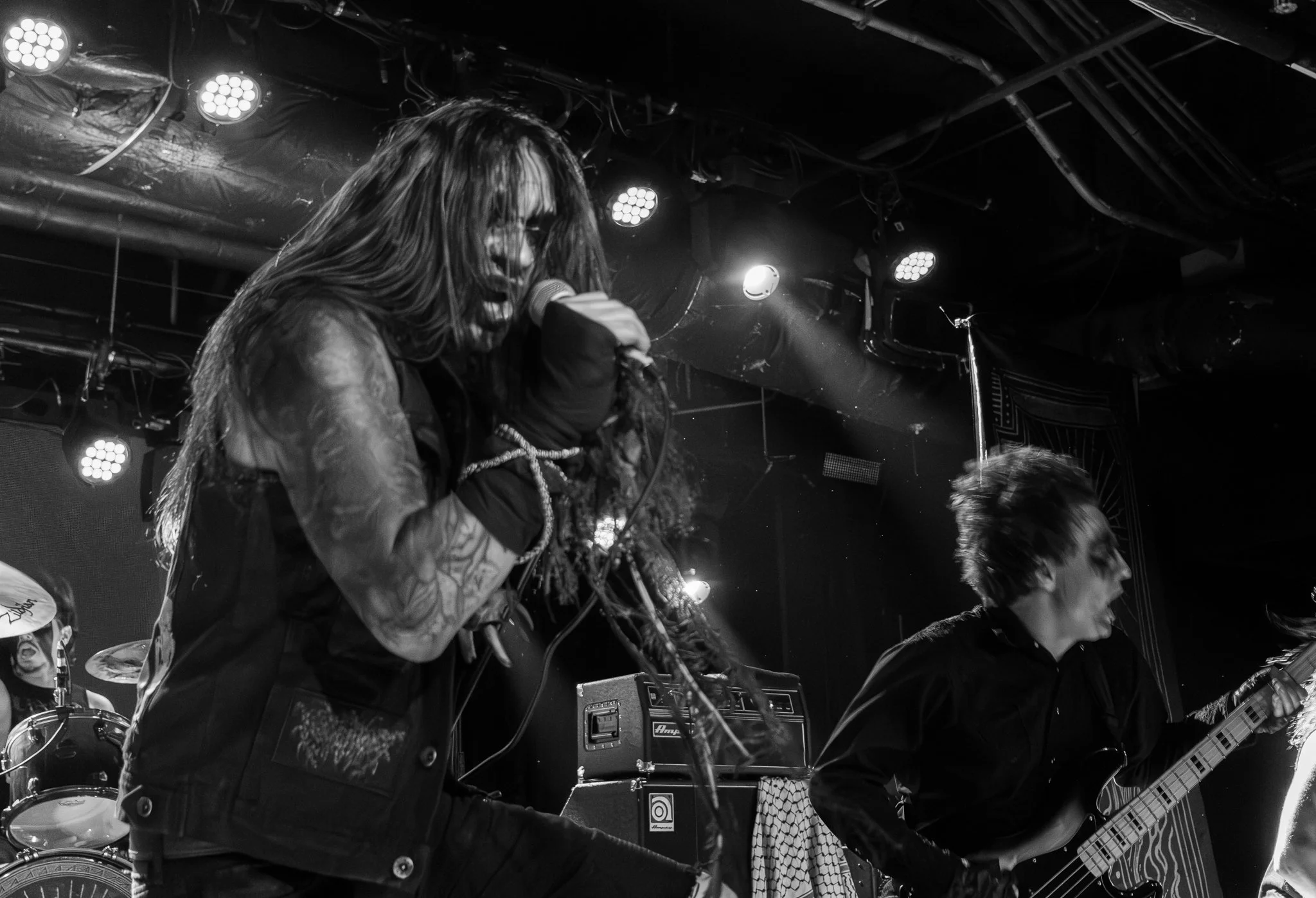 Black and white photo of a band performing on stage, singer with long hair singing into microphone, guitarist with messy hair playing guitar.