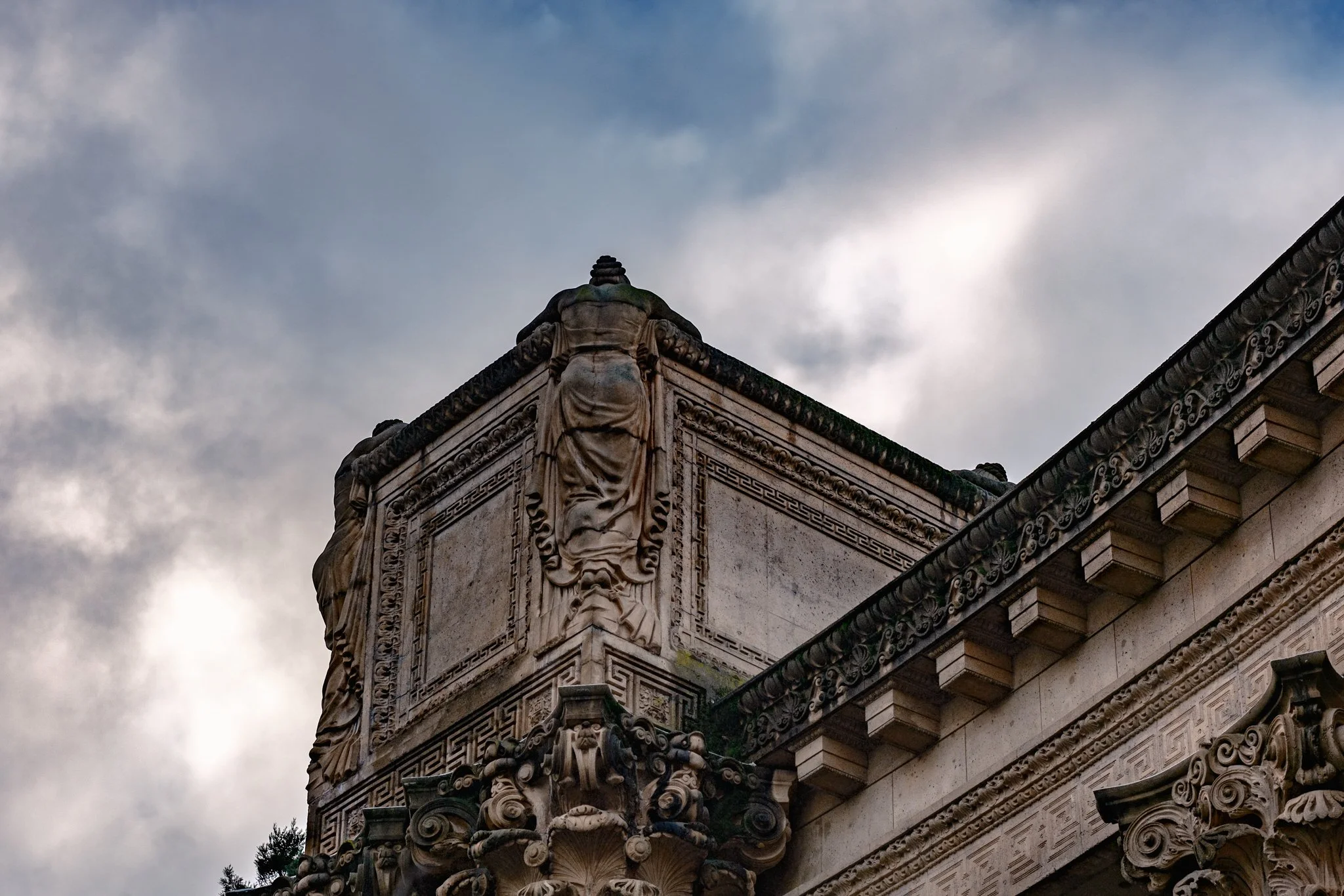 Close-up of an ornate, classical-style building corner with decorative stone carvings, sculptures, and intricate moldings, under a cloudy sky.