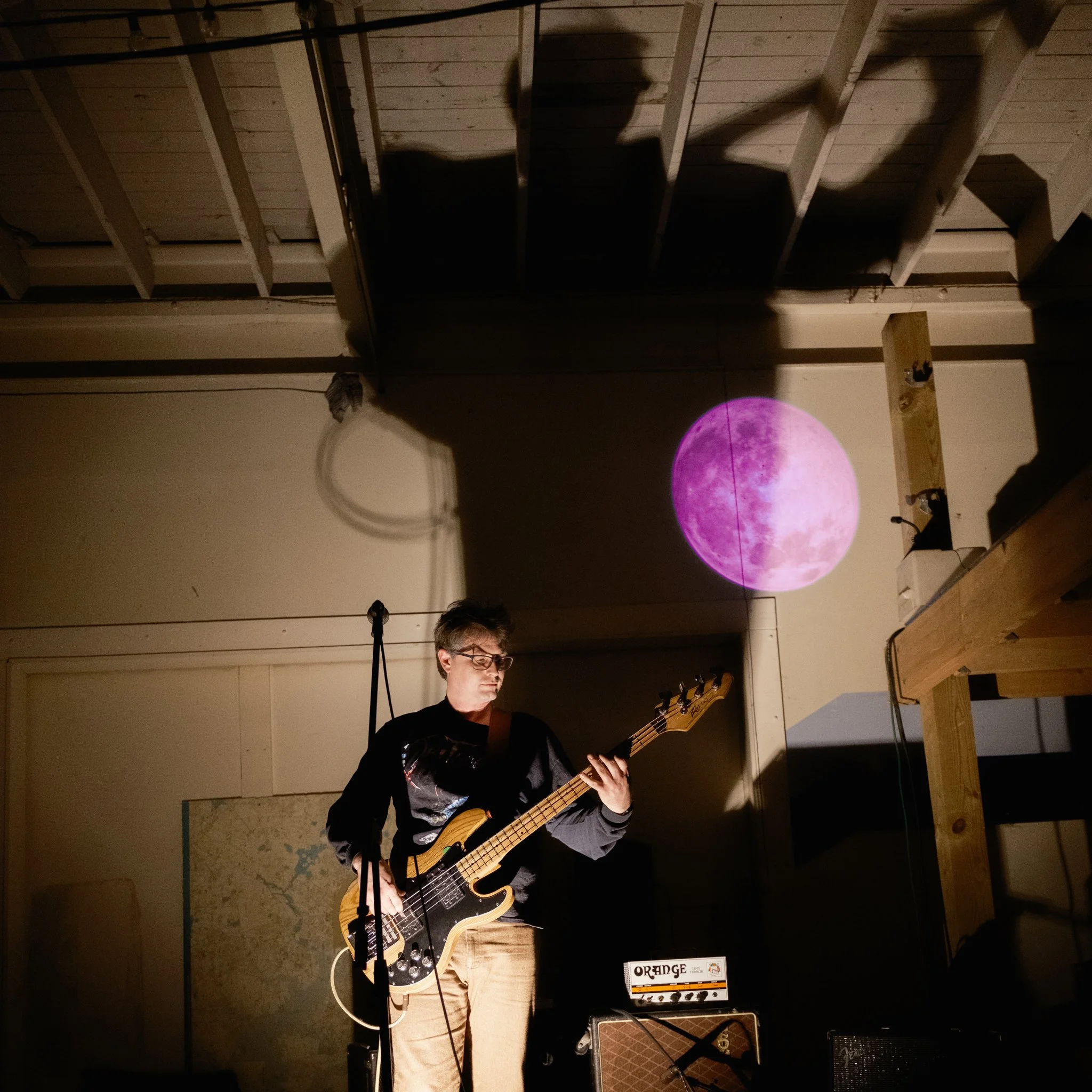 A person playing a bass guitar in a dimly lit room with a pink moon projected on the wall behind them.