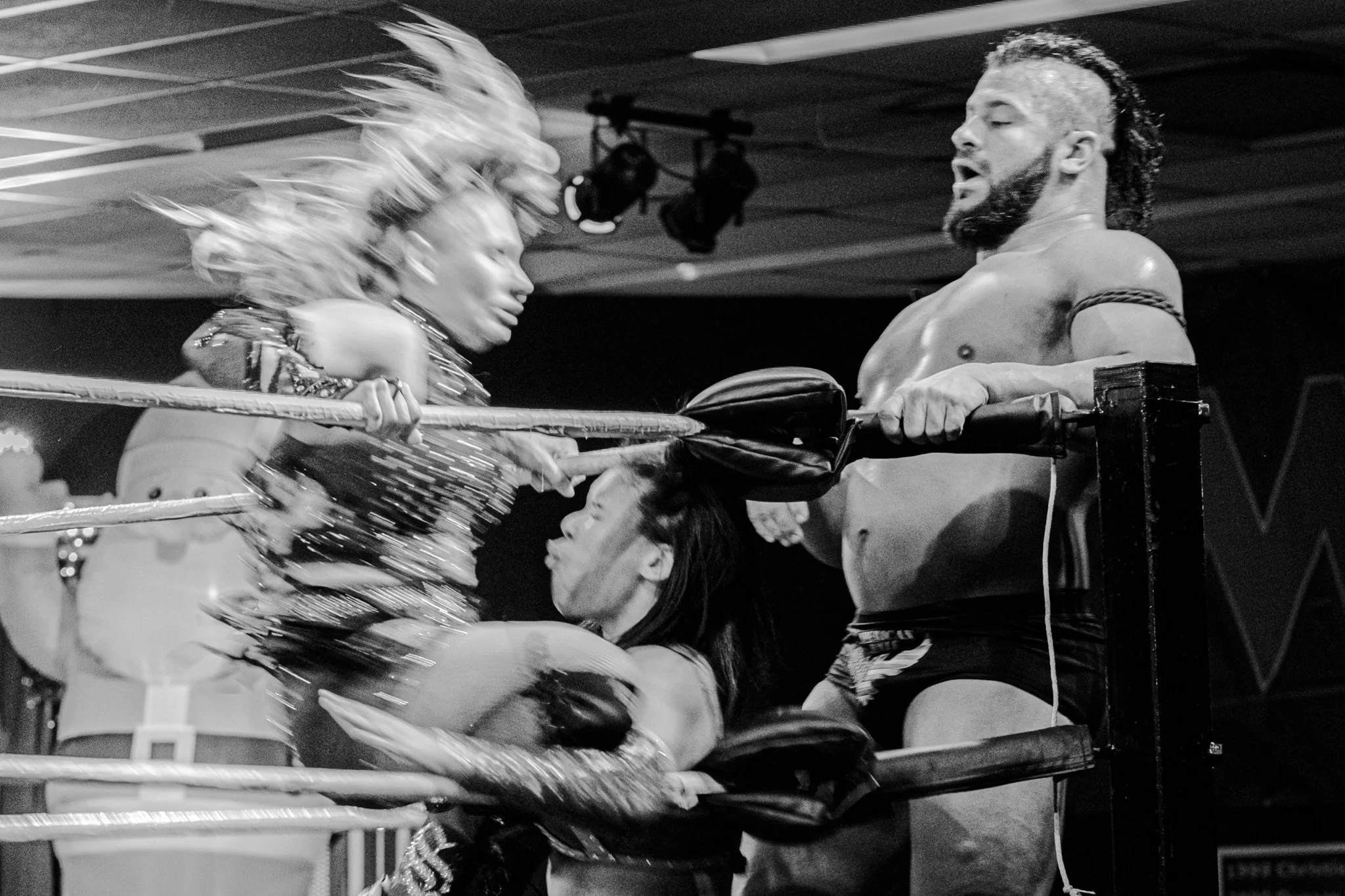 Two female wrestlers fighting inside a wrestling ring, with a male wrestler standing on the turnbuckle, holding the ropes and observing.