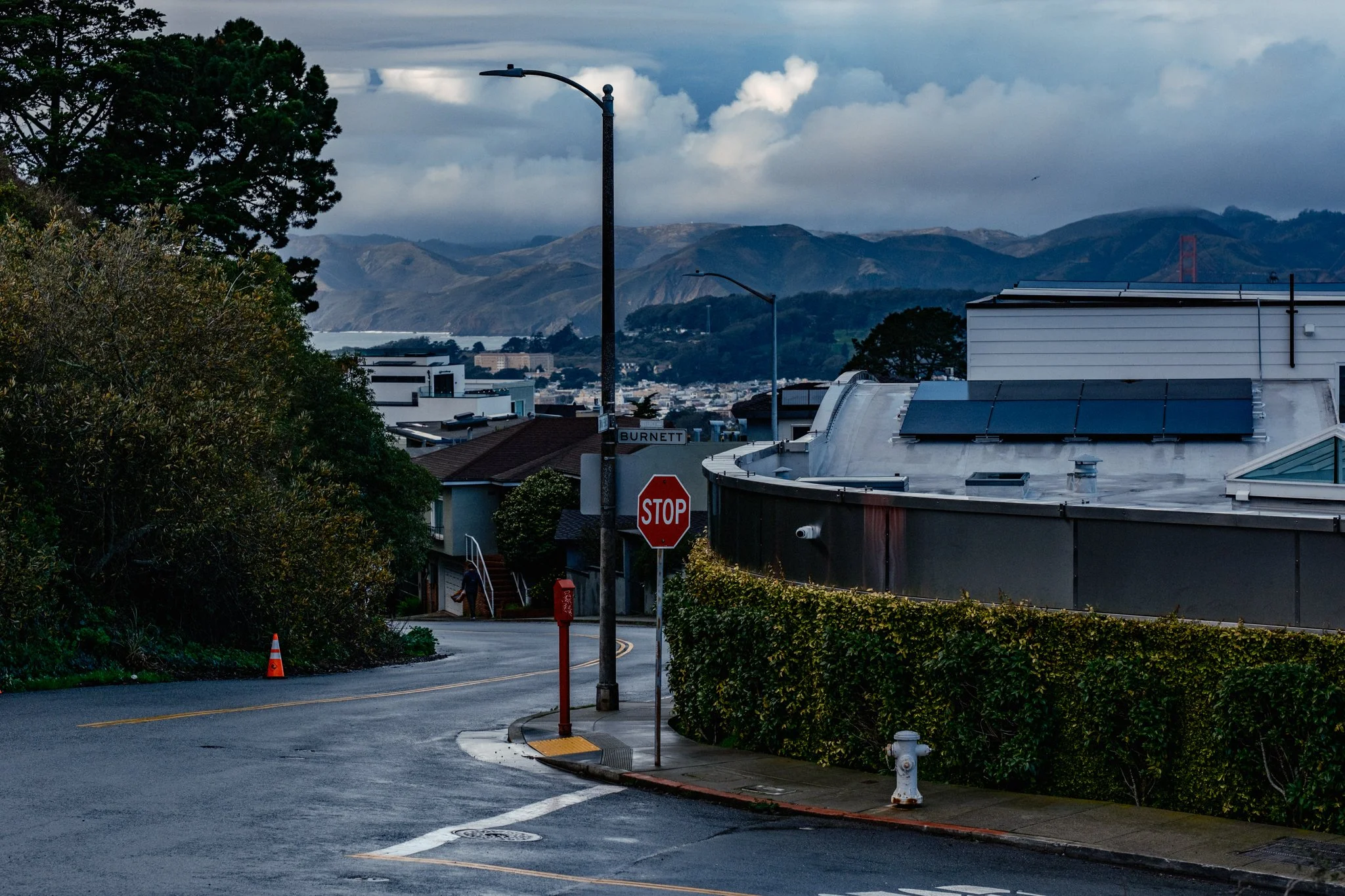A street corner with stop sign, street lights, and buildings with a mountainous landscape in the background on a cloudy day.