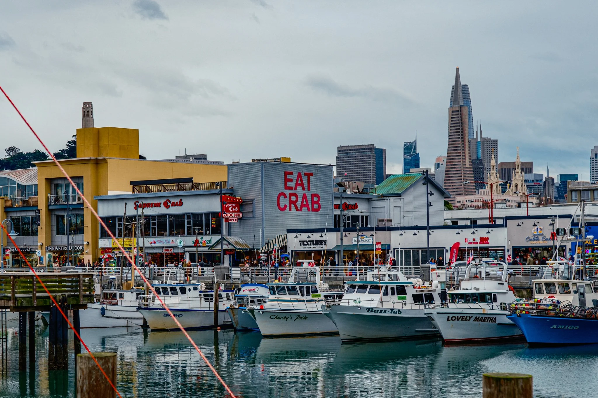 A marina with boats docked in the water in front of a commercial area with signs that say 'Eat Crab' and 'Supreme Crab.' In the background, a city skyline with tall skyscrapers, including one with a spire, is visible under a cloudy sky.