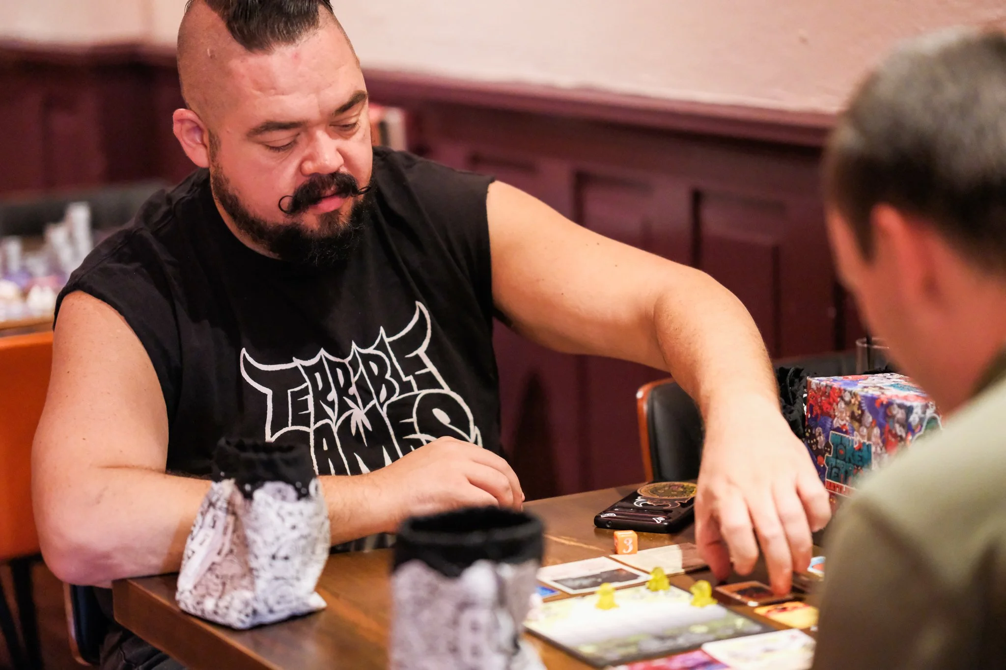 Two men are playing a board game at a table, with one man wearing a black sleeveless shirt with a graphic design, and the other wearing glasses and a beige shirt. The table has game pieces, cards, a dice, and a wrapped gift.