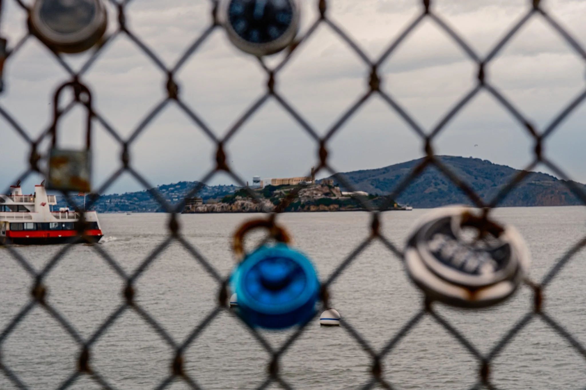 A view of Alcatraz Island through a chain-link fence with padlocks attached, overlooking San Francisco Bay and a ferry boat sailing nearby.