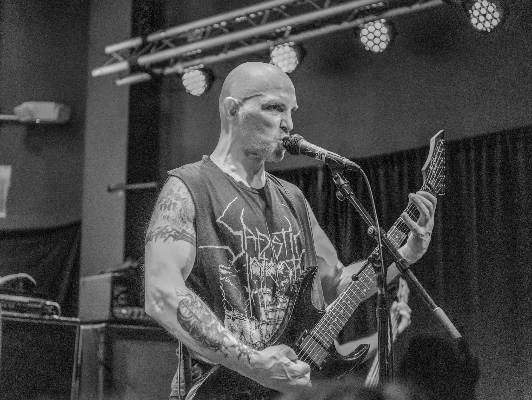 A bald man with tattoos on both arms sings into a microphone and plays an electric guitar on stage, wearing a sleeveless T-shirt with a graphic design.