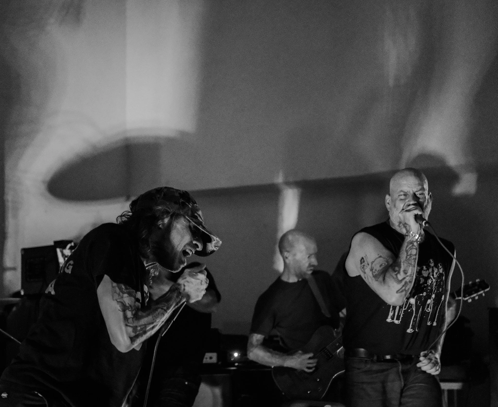 Black and white photo of three band members performing live: a singer with tattoos holding a microphone, a guitarist, and another musician in the background.