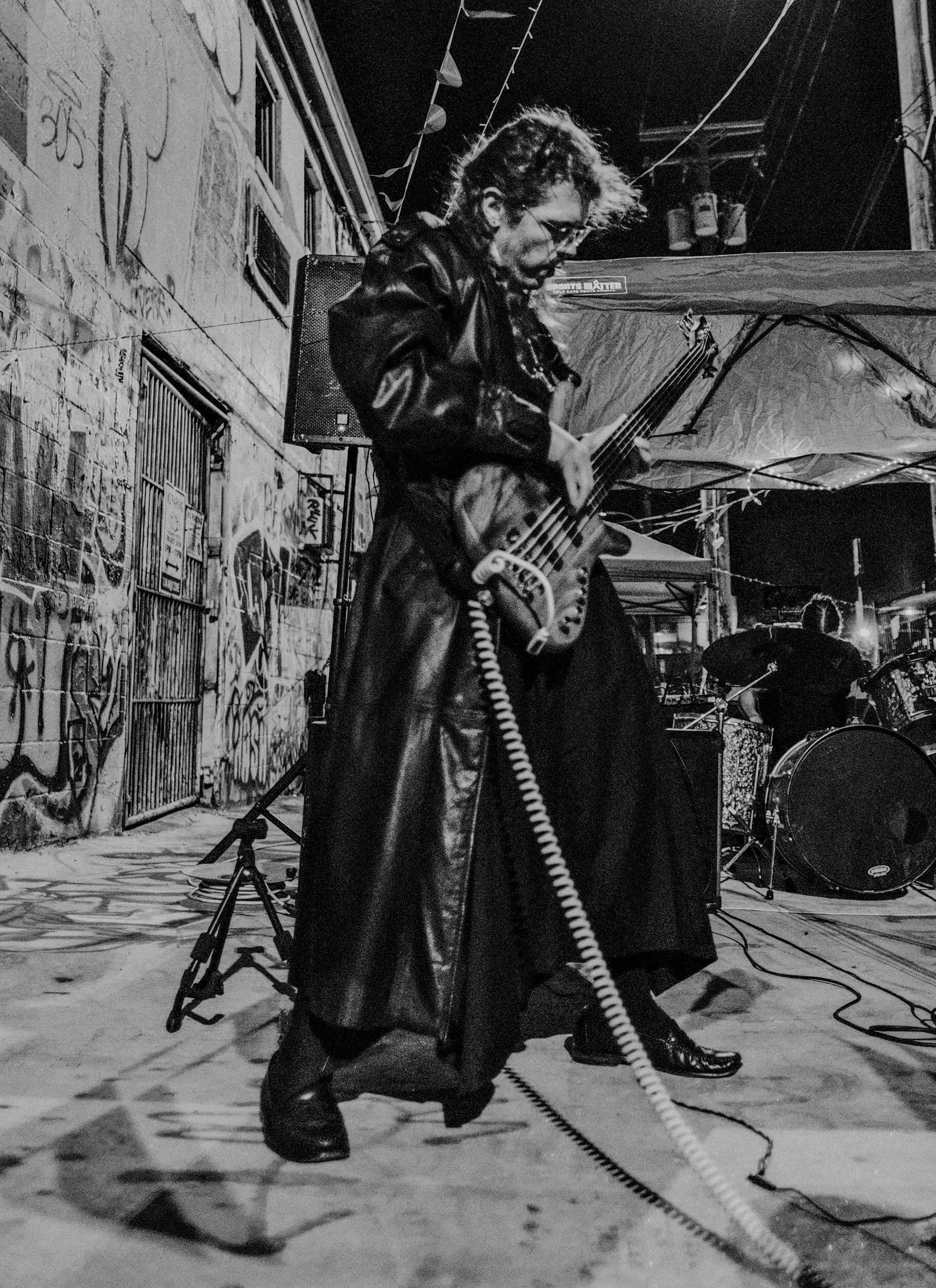 A musician playing an electric bass guitar during a night outdoor performance, with graffiti-covered brick wall in the background and another band member on drums.