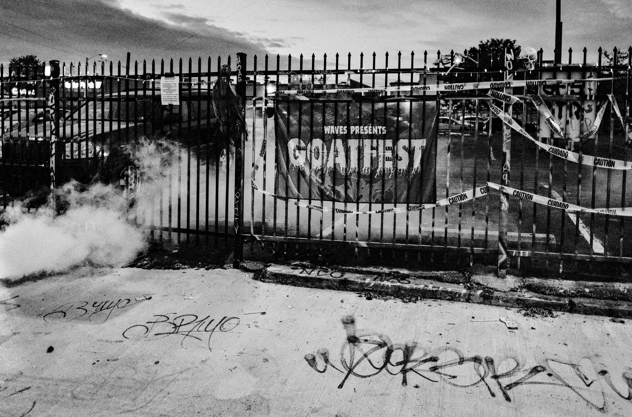 A black and white photo of a metal fence decorated with caution tape and a banner that reads "CIRCUS" with a spooky design. There is graffiti on the ground in front of the fence, and smoke or fog on the left side of the image. The sky is cloudy, and 
