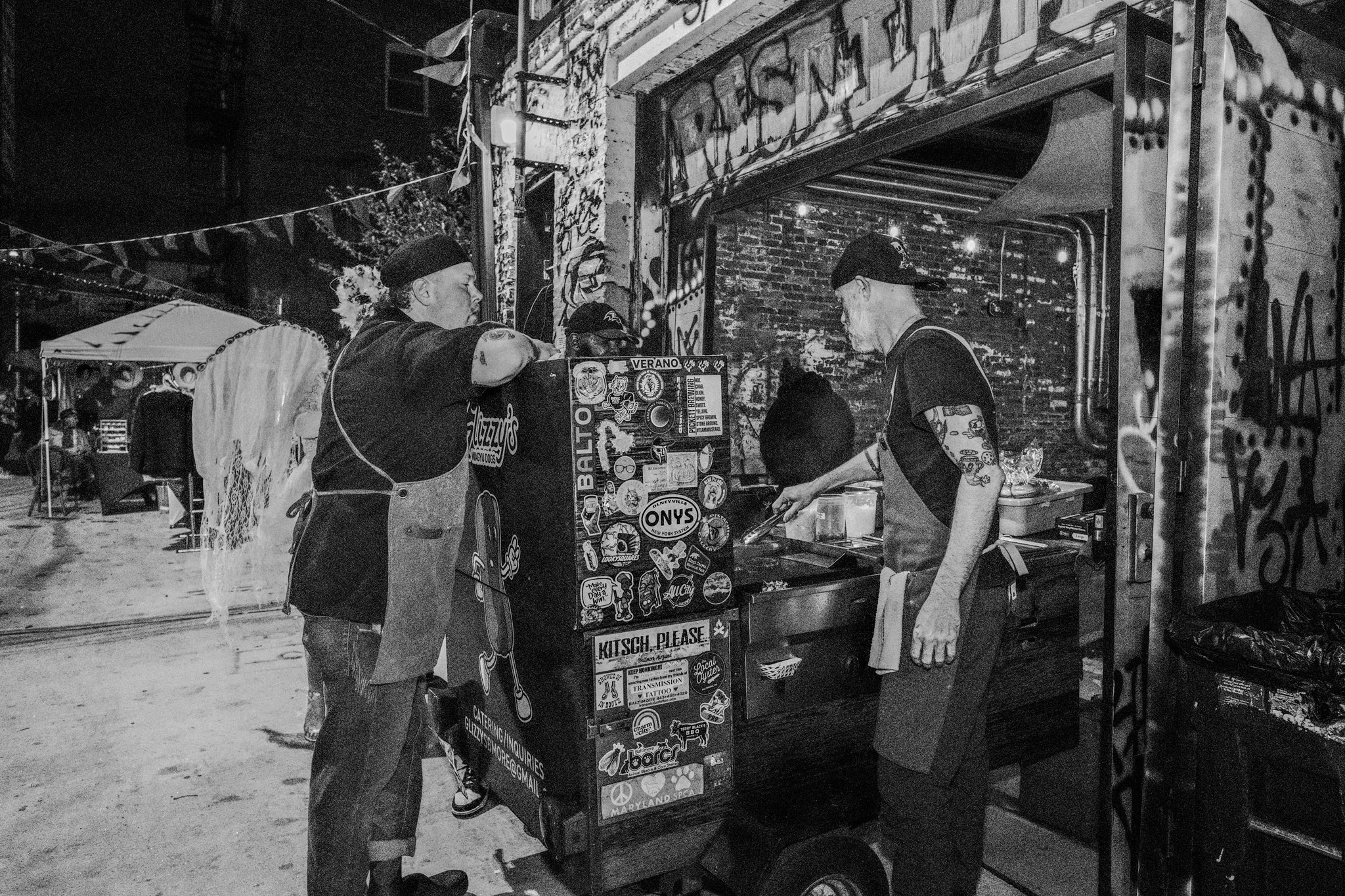 Night scene of a street food stand with two vendors preparing food and a customer ordering. The stand is decorated with stickers and graffiti, with people in the background under string lights.