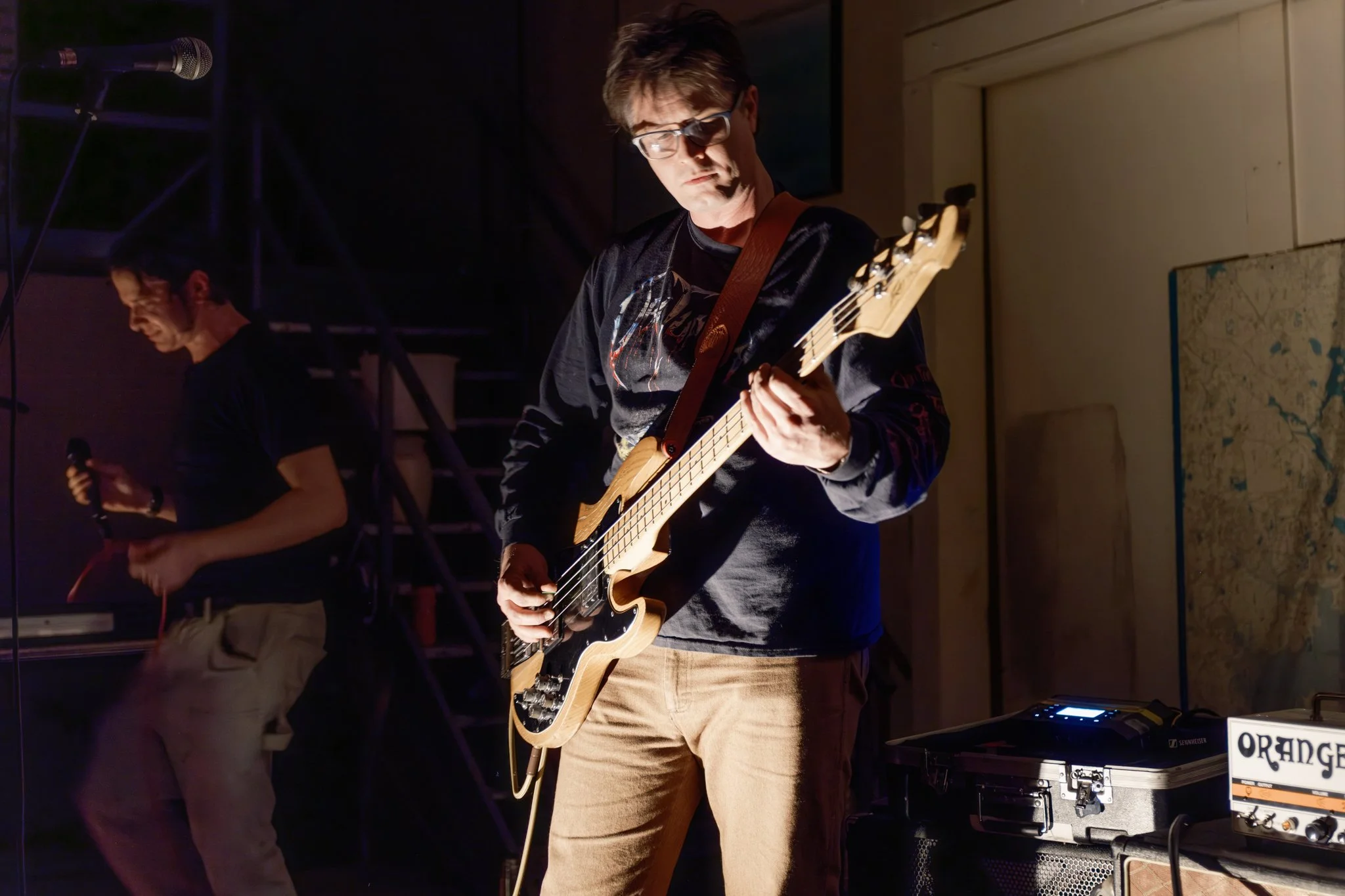 Musician playing electric guitar on stage, wearing glasses, black sweatshirt, and khaki pants, with another person holding a microphone in the background.