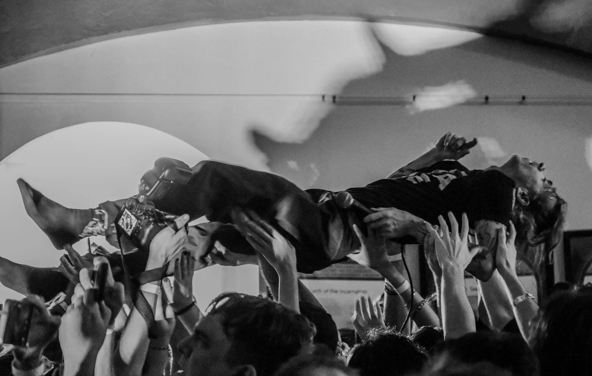 A person being carried above a crowd of people, with many hands supporting them, indoors with a ceiling and a light source in the background.