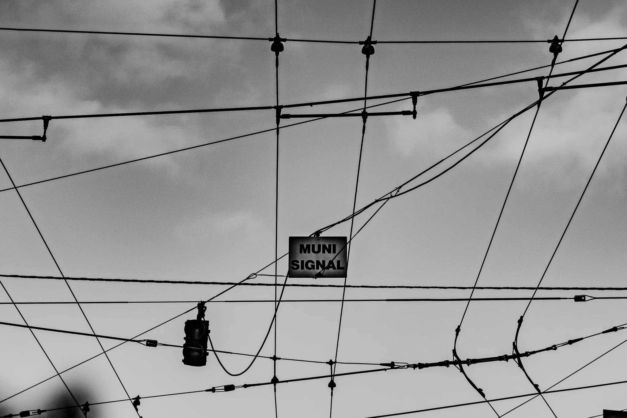 Overhead view of electrical wires and streetlights against a cloudy sky, with a sign reading 'MUNI SIGNAL' hanging among the wires.