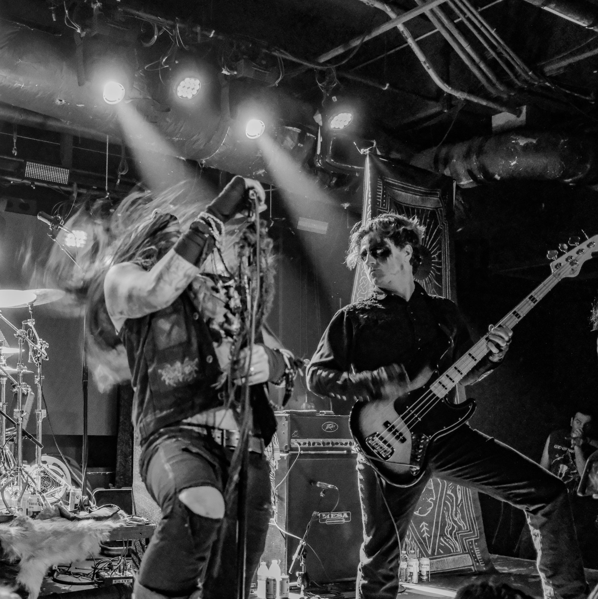 A black and white photo of a live rock band performing on stage, with two musicians at the forefront: a singer with long hair, wearing ripped jeans, and a bassist with short hair and dark makeup, playing an electric bass guitar.