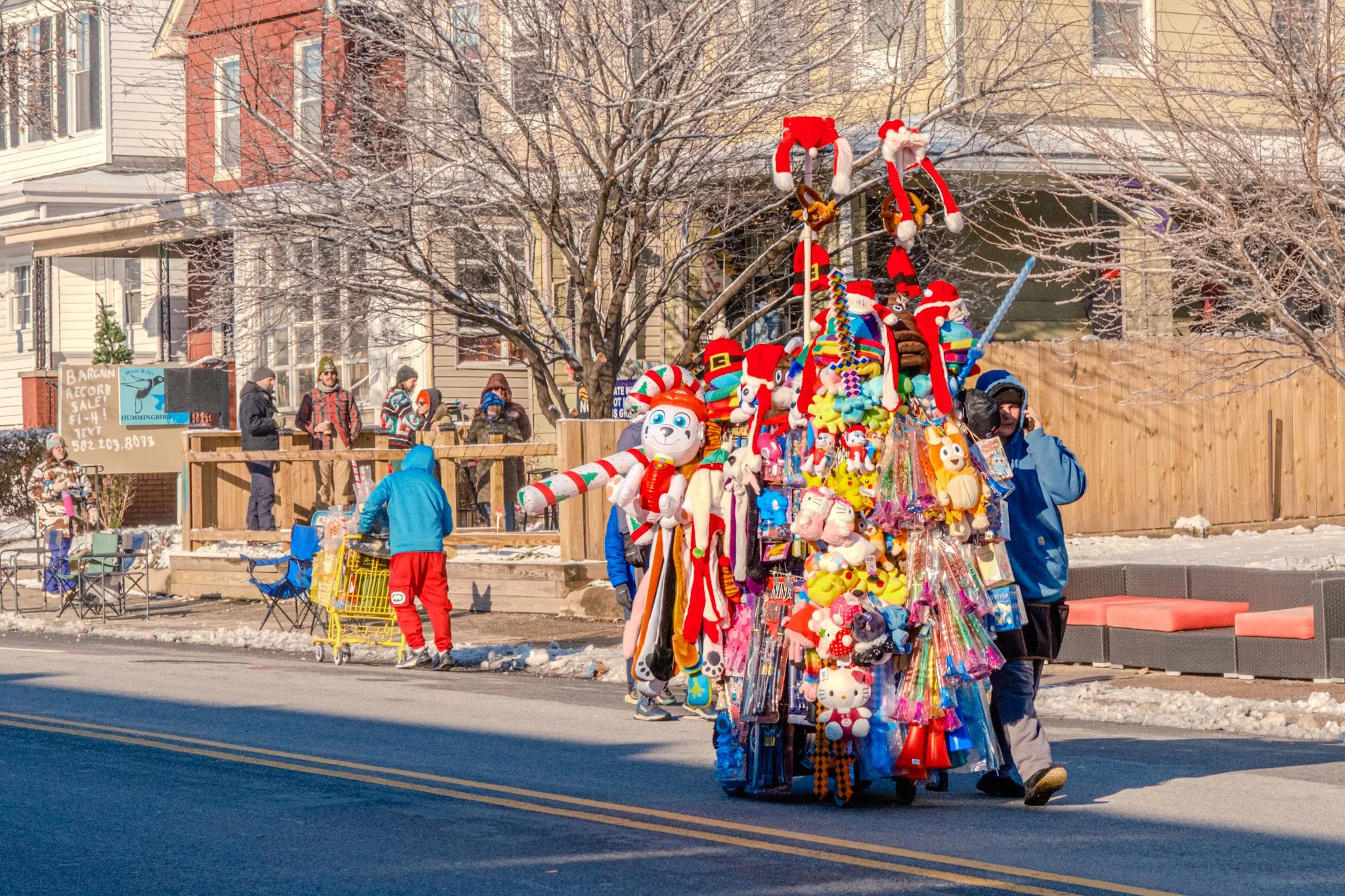 A street scene during winter with a Christmas-themed float decorated with plush toys and Christmas ornaments. People are seen walking and standing along the sidewalk, bundled in winter clothing. Snow covers parts of the ground and a tall, leafless tr