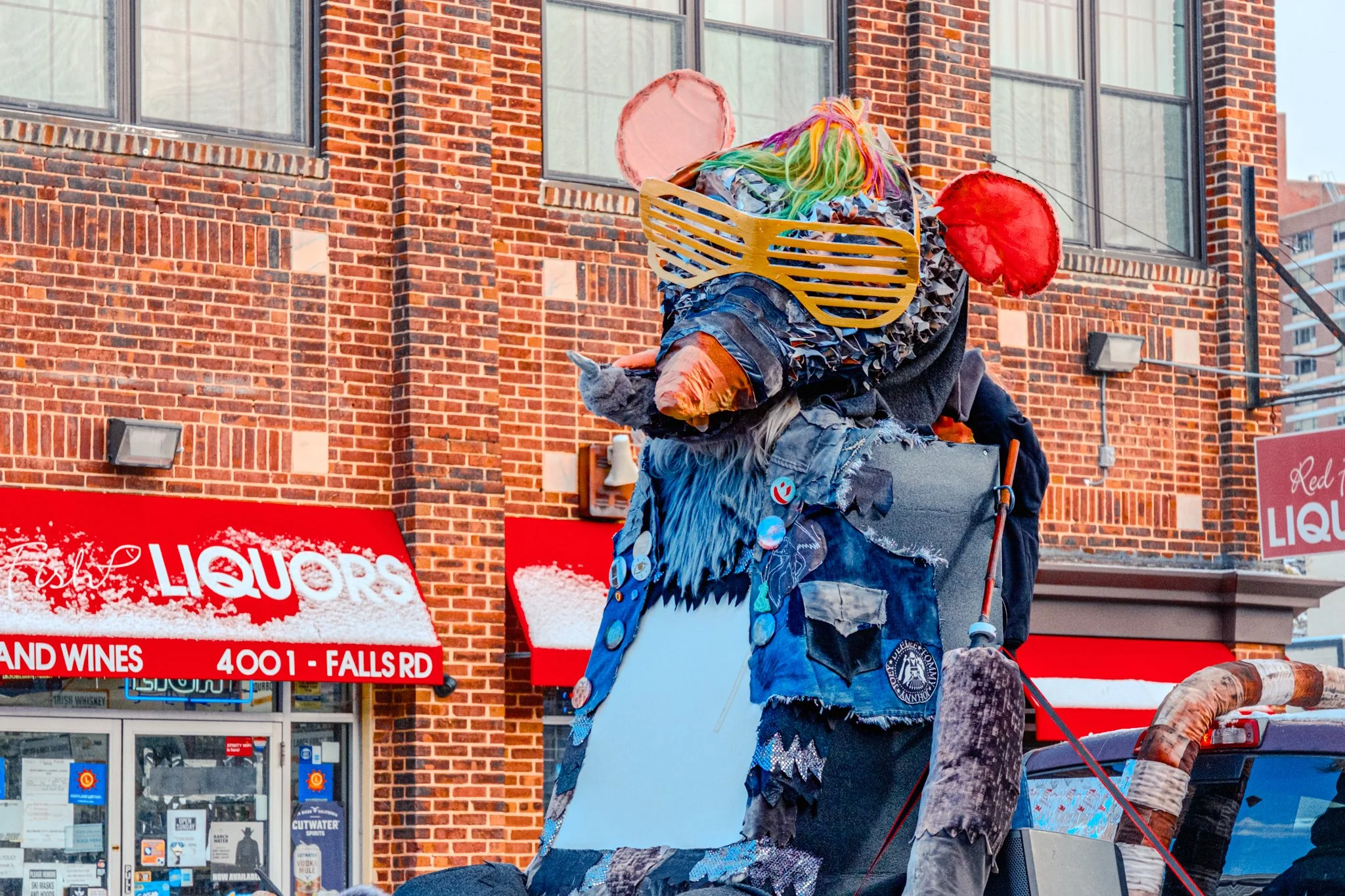 Large colorful puppet or sculpture resembling an animal or mascot, with a large mask and bright rainbow hair, sitting on a vehicle in an urban area with brick buildings and store signs in the background.