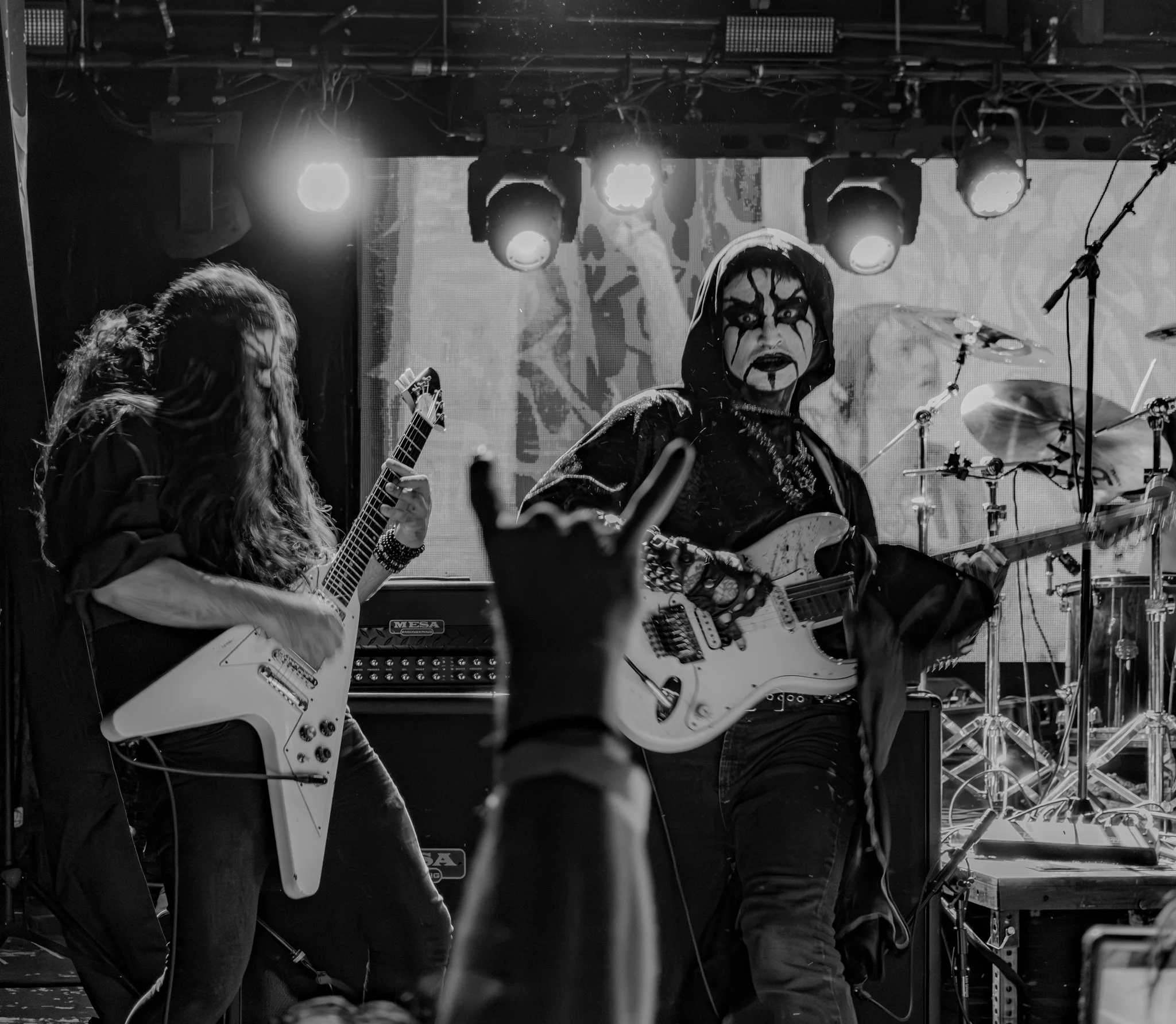 A black and white photo of a live music performance on stage, featuring two guitarists and a drummer. One guitarist on the right has face paint, a hood, and long dark hair, playing an electric guitar. The other guitarist on the left has long wavy hai