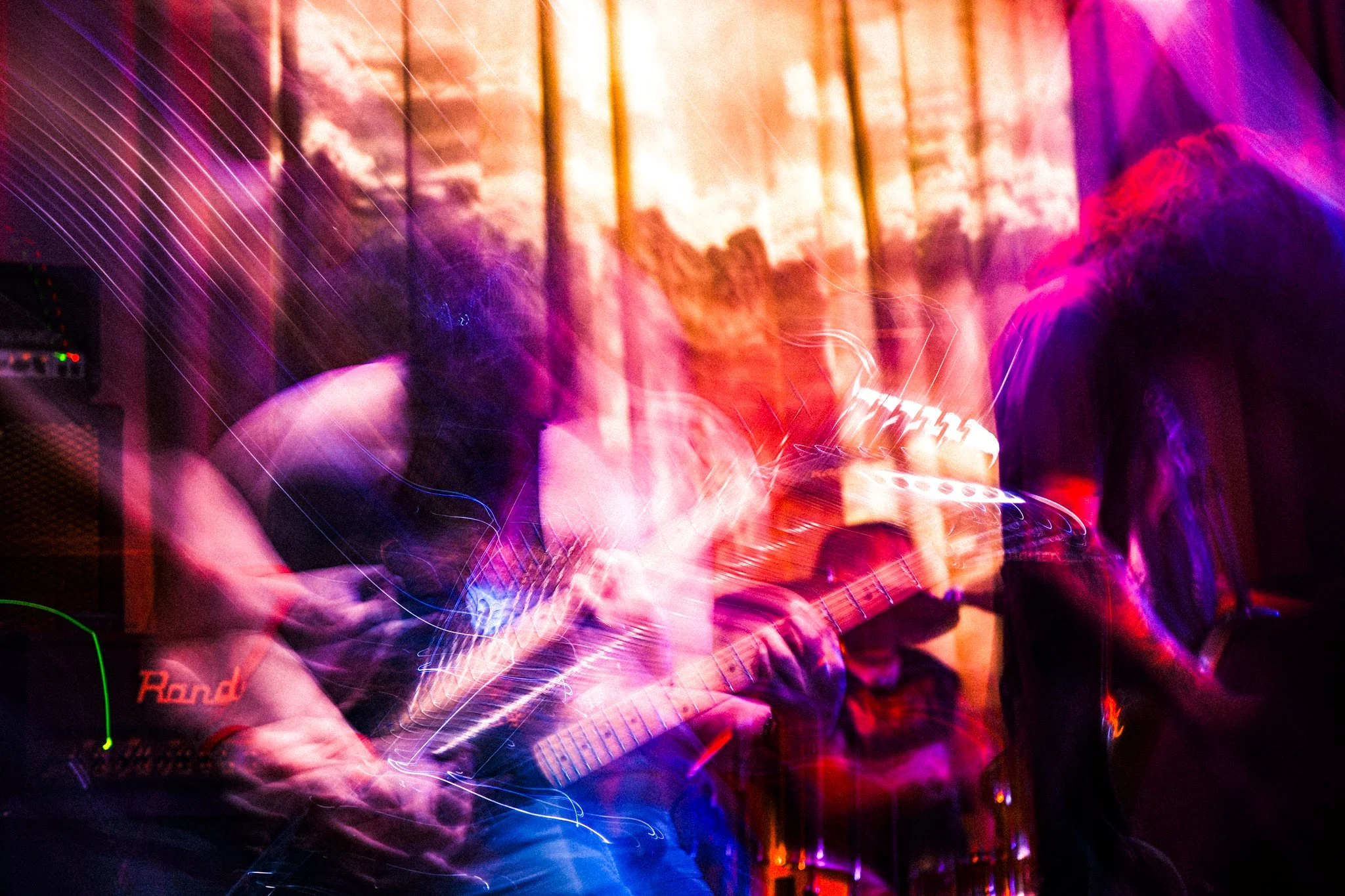Two musicians playing guitars on stage in a dimly lit venue with purple and red lighting, and a blurred, dynamic motion effect.