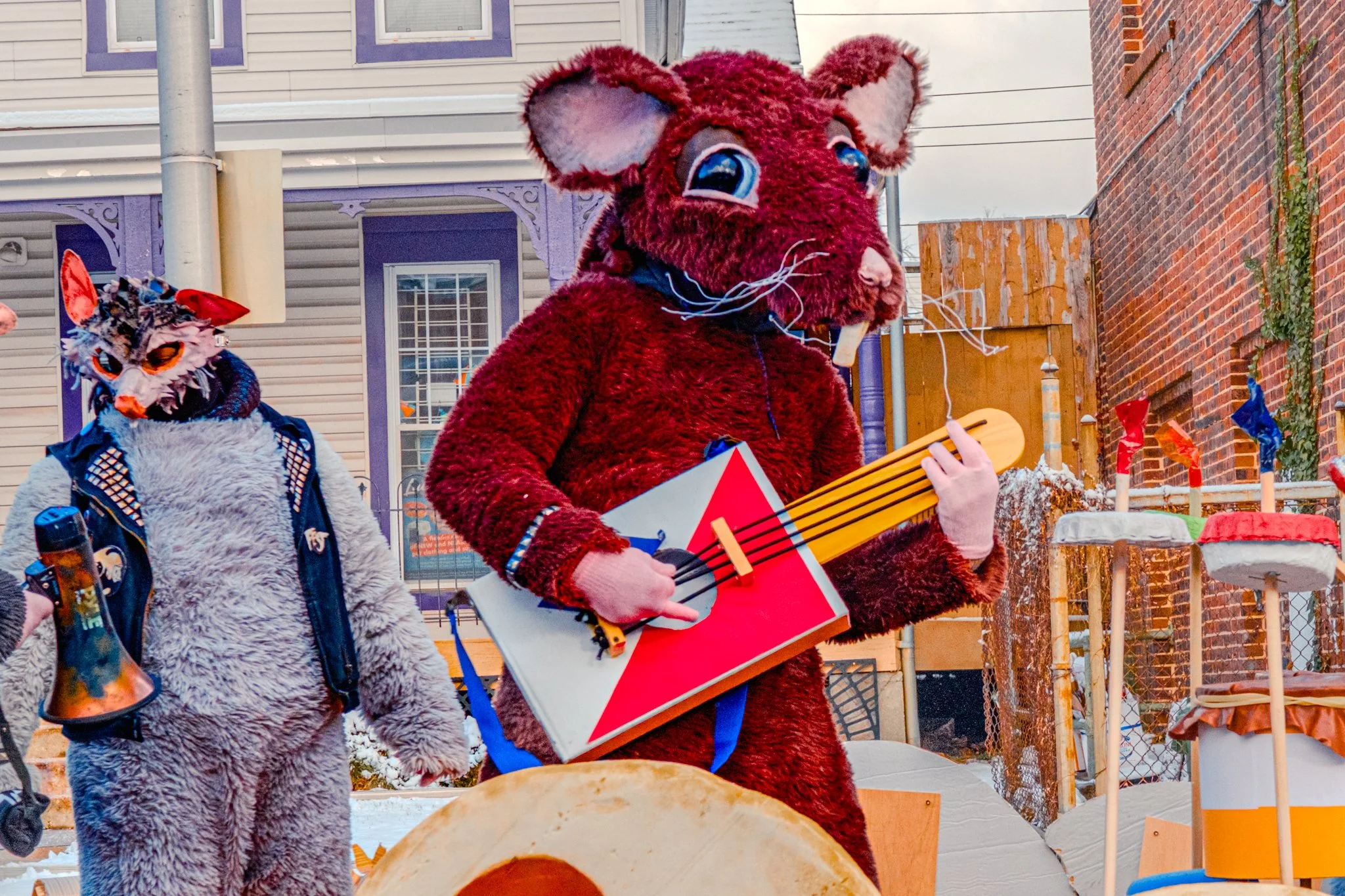 People dressed in colorful animal costumes, including a mouse with a guitar, participating in a parade or outdoor event with decorated structures and buildings in the background.