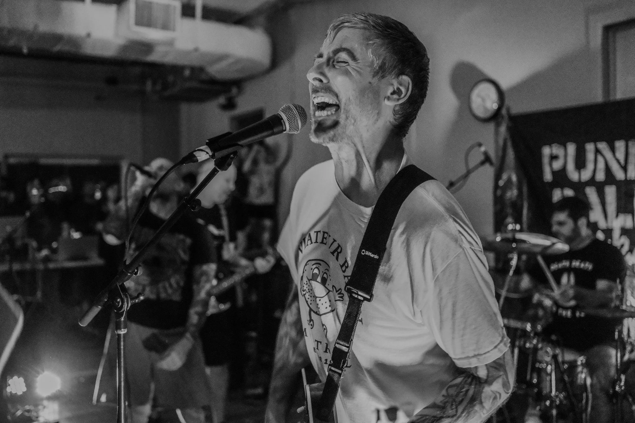 A male singer with tattoos on his arm, singing passionately into a microphone while playing guitar during a live performance, with band members in the background.