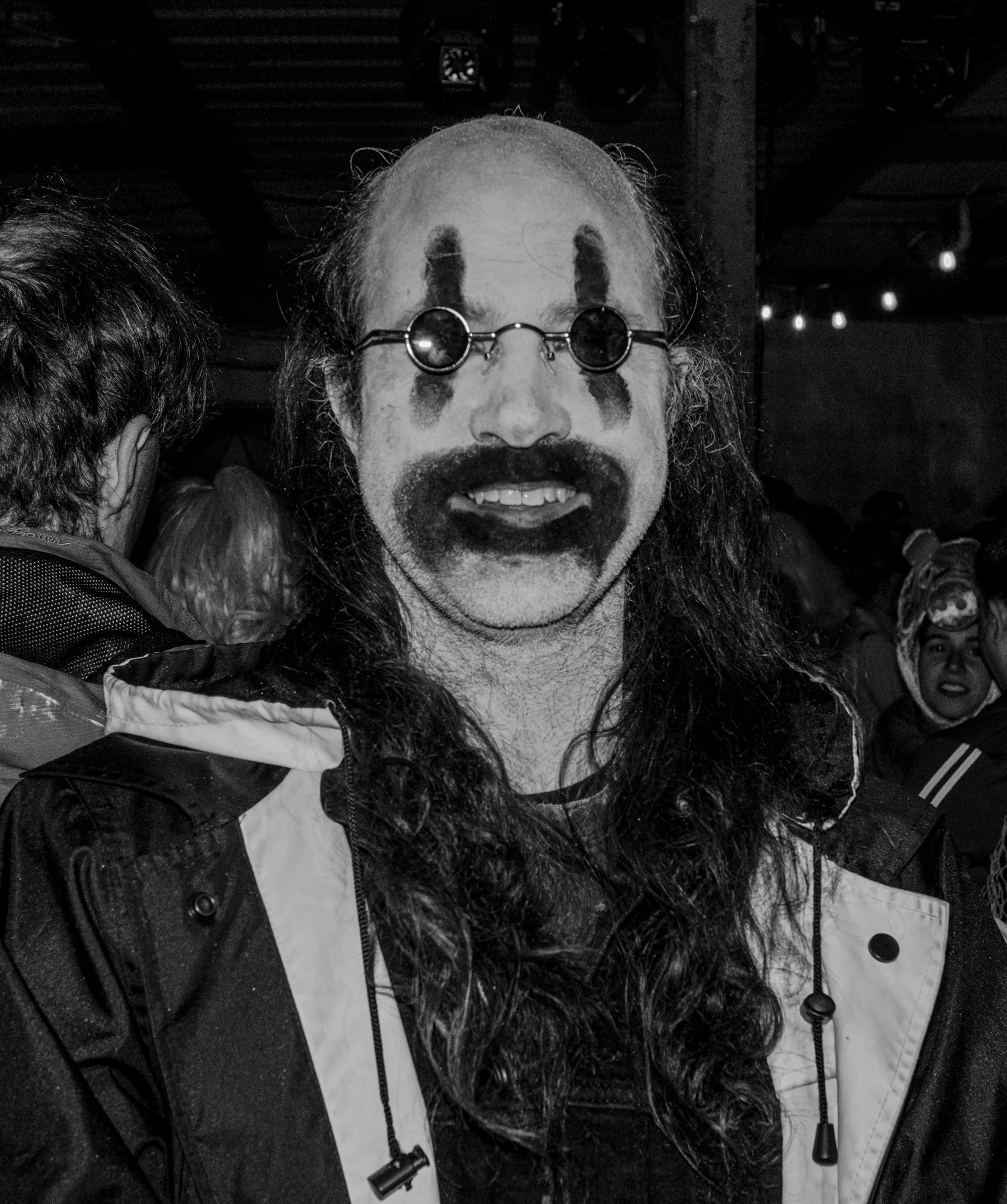 Person dressed as a clown with face paint, sunglasses, and long hair, smiling at a crowded event.