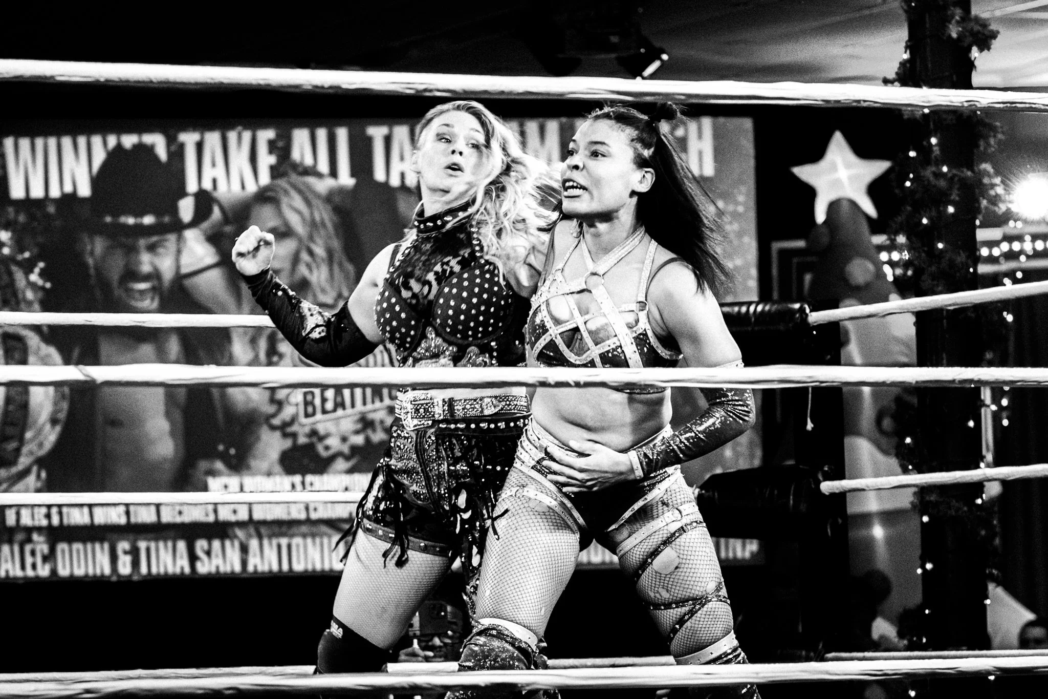 Two female wrestlers in action inside a wrestling ring. One is wearing a patterned top and shorts, the other in a strappy crop top and shorts, both in a confrontation. A large banner and festive decorations are visible in the background.