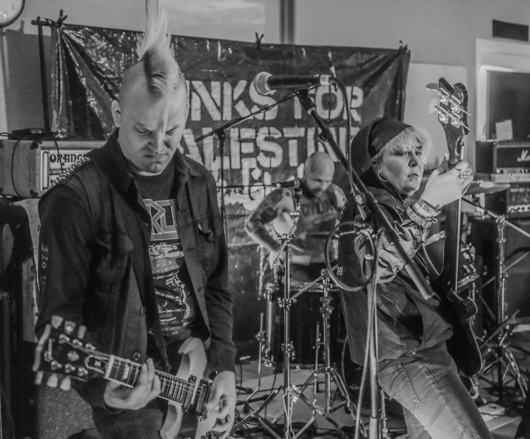 A black and white photo of a band performing live on stage. There are two guitarists in the foreground, one male with a mohawk hairstyle and another female with short hair, wearing a hooded jacket, holding an electric guitar. In the background, a dru