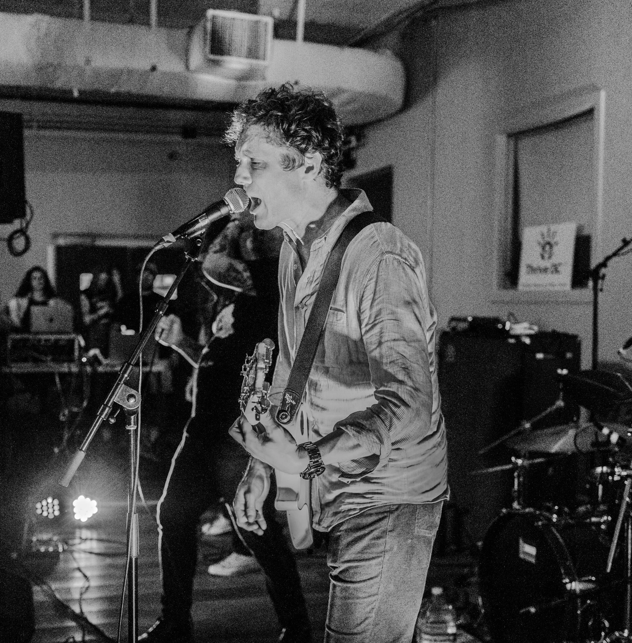Black and white photo of a musician singing into a microphone and playing an electric guitar, with a drummer in the background and a woman at a computer to the left.