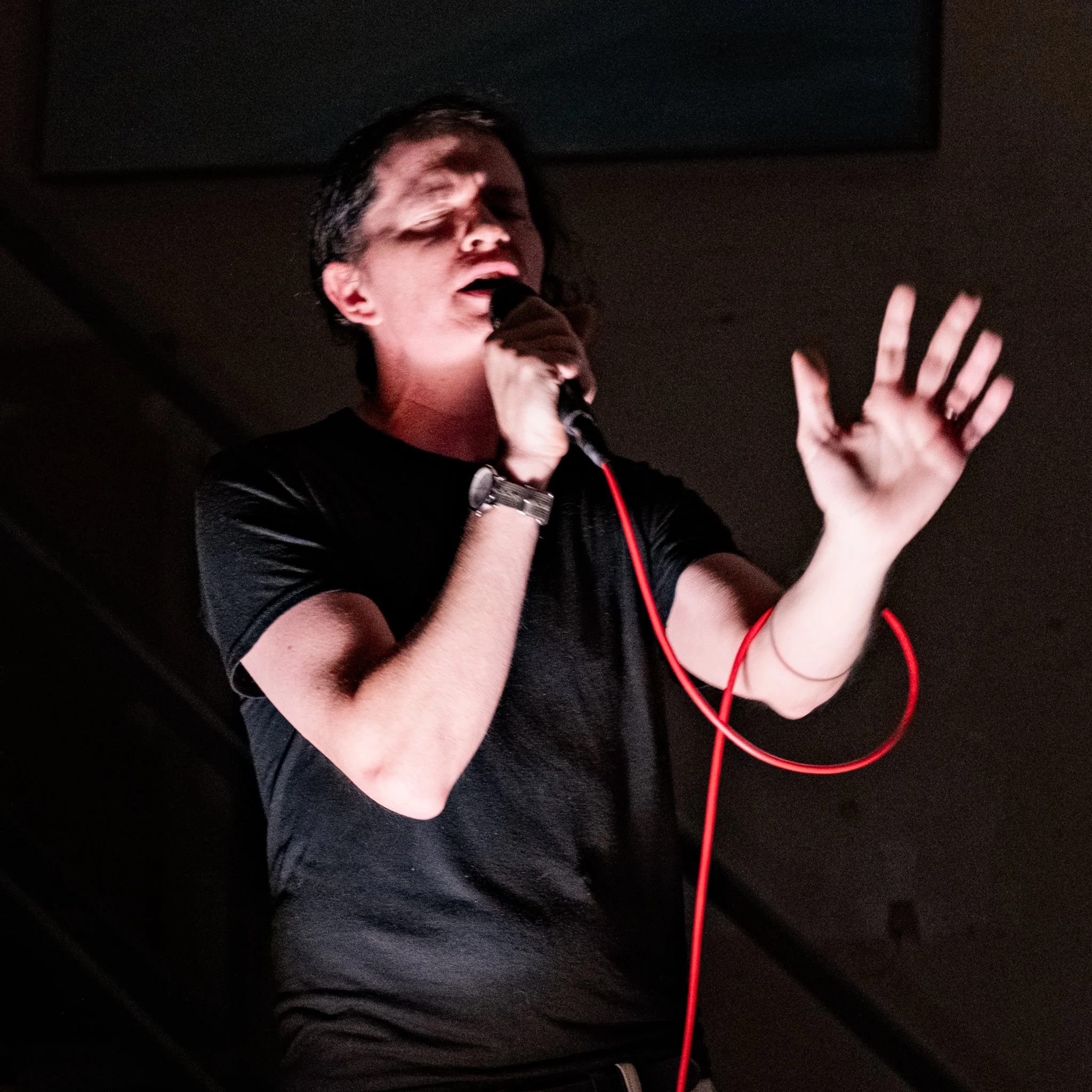 A person singing into a microphone with eyes closed, wearing a black shirt, a watch, and a red cord attached to the microphone.
