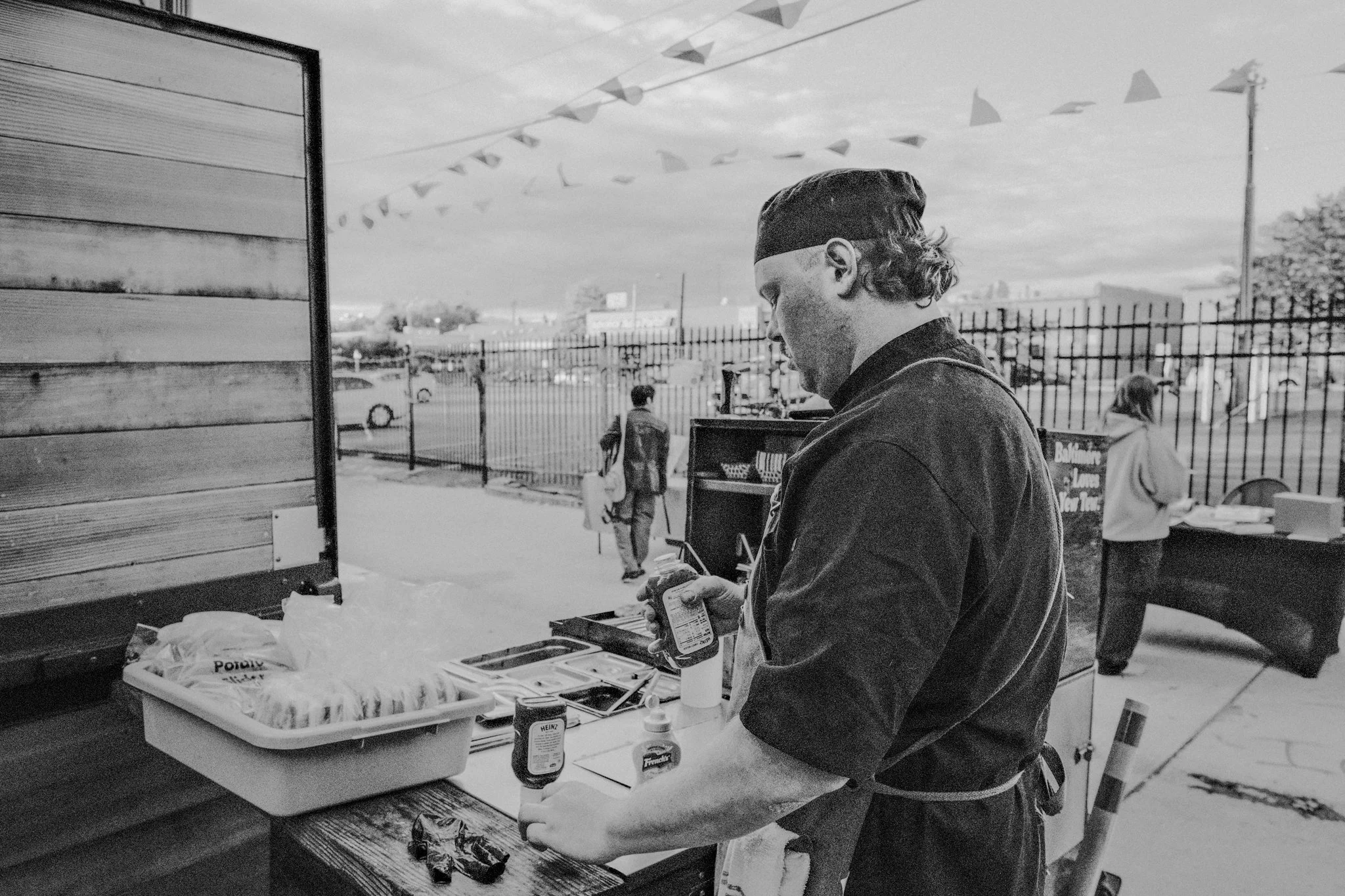A man preparing food at an outdoor food stand, wearing a apron and a hat, with condiments and food items on the counter. Other people are in the background near a fence and street.