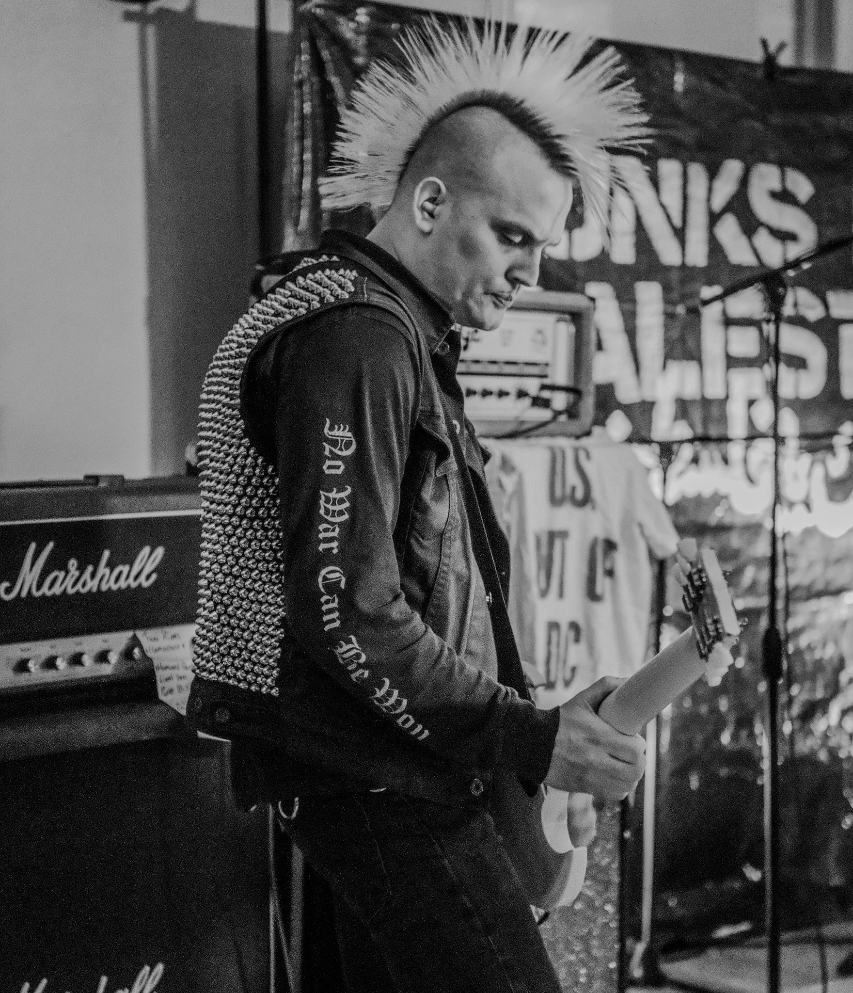 A person with a mohawk hairstyle reading a rolled-up paper in a music practice space, with Marshall amplifier and banners in the background.