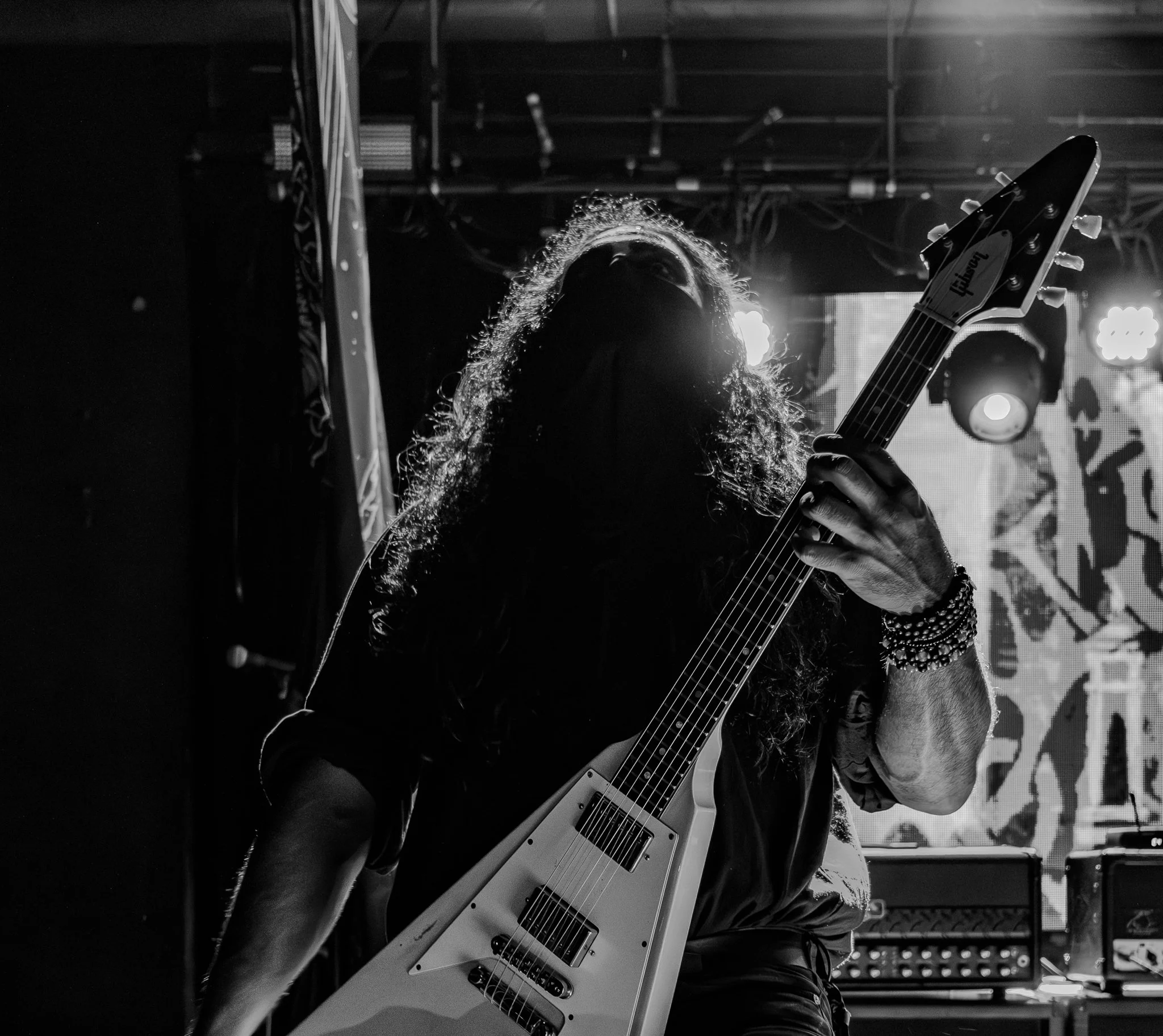 A musician with long curly hair playing a white electric guitar on stage, backlit by bright lights, in black and white.