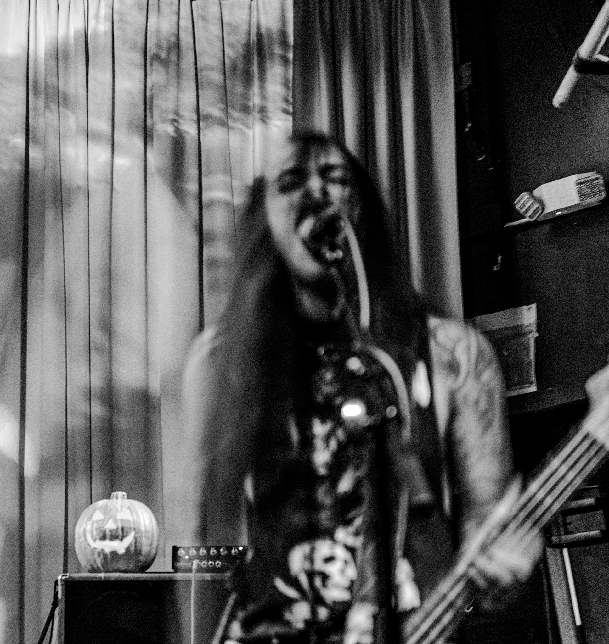 A person with long hair playing a bass guitar, wearing a skeleton-themed shirt, singing or shouting with a microphone in a small, dimly lit room decorated with a pumpkin and other items.