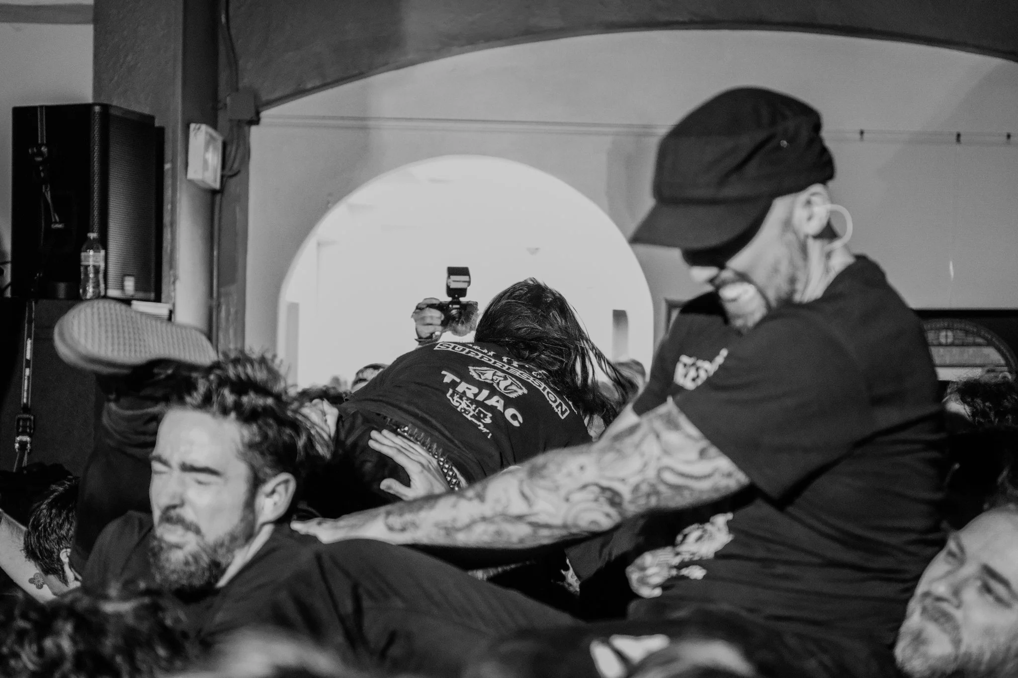 People participating in a wrestling match or fight, with one person in black clothing and another with tattoos and glasses, in a crowded indoor setting.