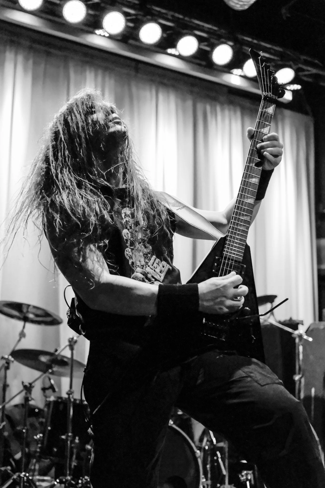 Black and white photo of a guitarist with long hair playing an electric guitar on stage, with drums in the background and stage lights overhead.