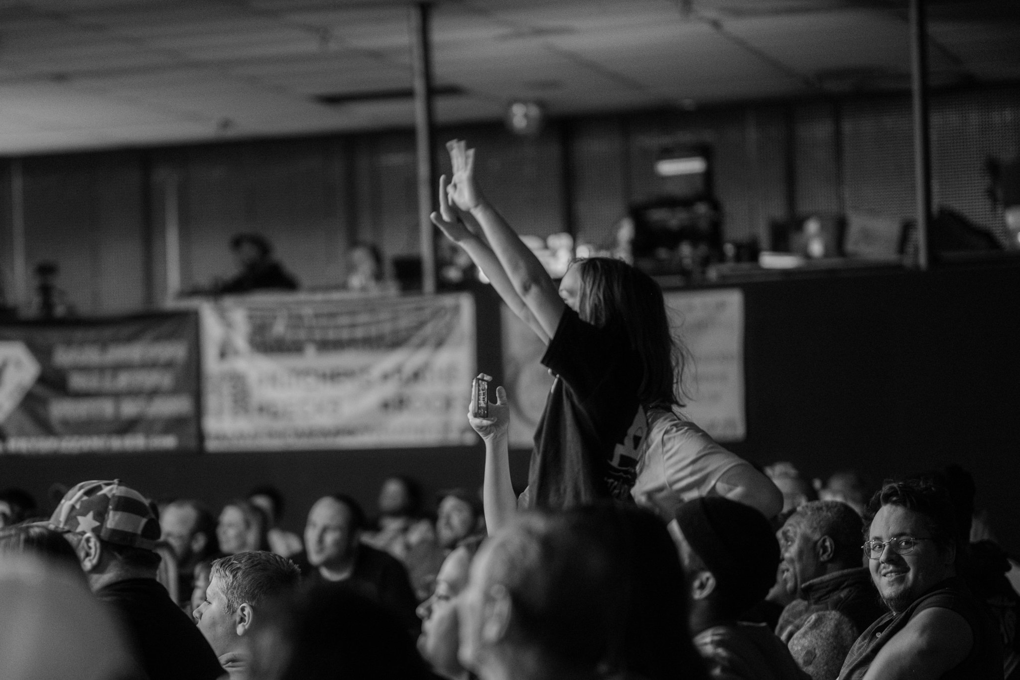 A woman standing in a crowd at an indoor event, holding a small object in her left hand, with her arms raised above her head, and a mask covering her face.