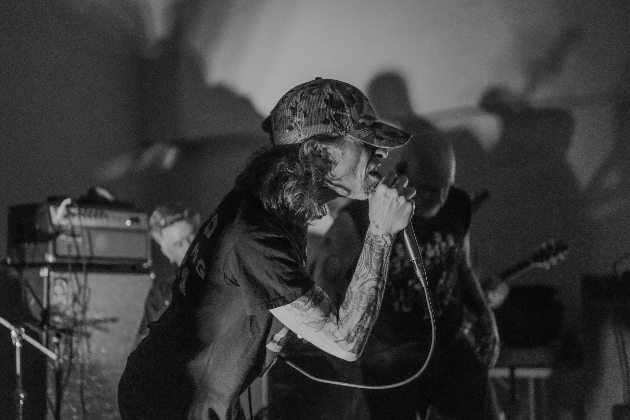 A black and white photo of a man with long hair, wearing a camouflage hat, singing into a microphone during a live music performance. Other band members and musical equipment are visible in the background.